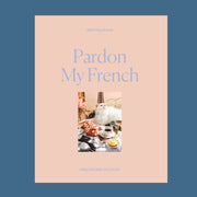 A peachy tan colored puzzle box with a 1000 piece puzzle inside with an image of a white fluffy cat surrounded by French goods such as croissants, macarons, champagne and the Eiffel Tower.