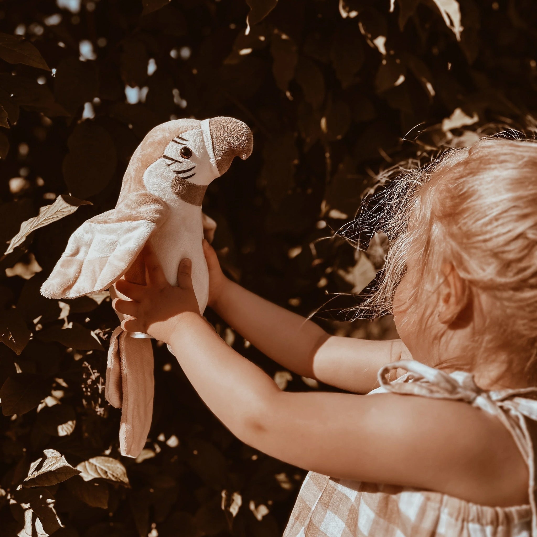 Child holding a stuffed parrot toy in front of a leafy background