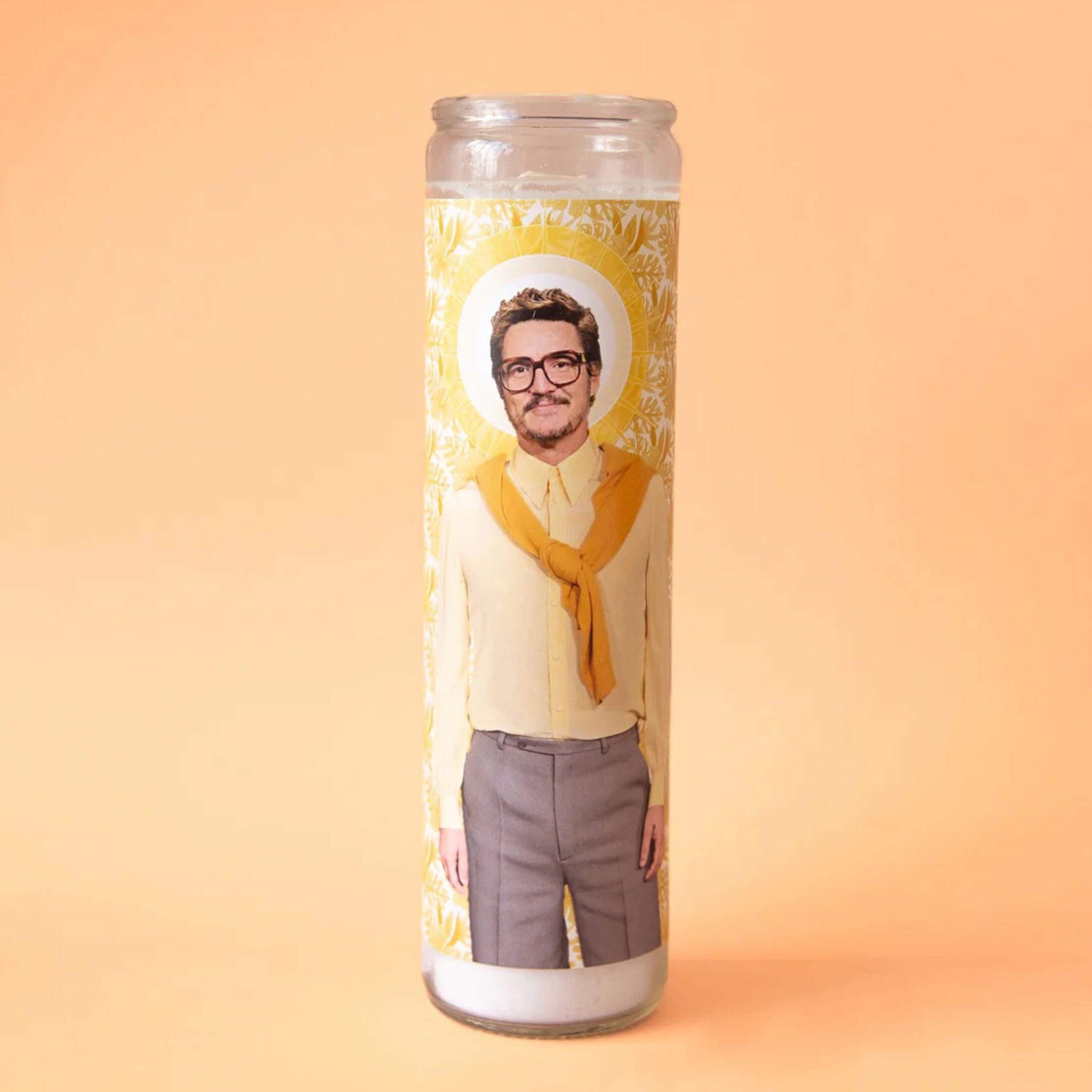 On a yellow background is a tall thin prayer candle in a clear glass jar with a label on the front featuring a photograph of Pedro Pascal in glasses and wearing grey slacks a yellow button down and a orange sweater tied around his shoulders.
