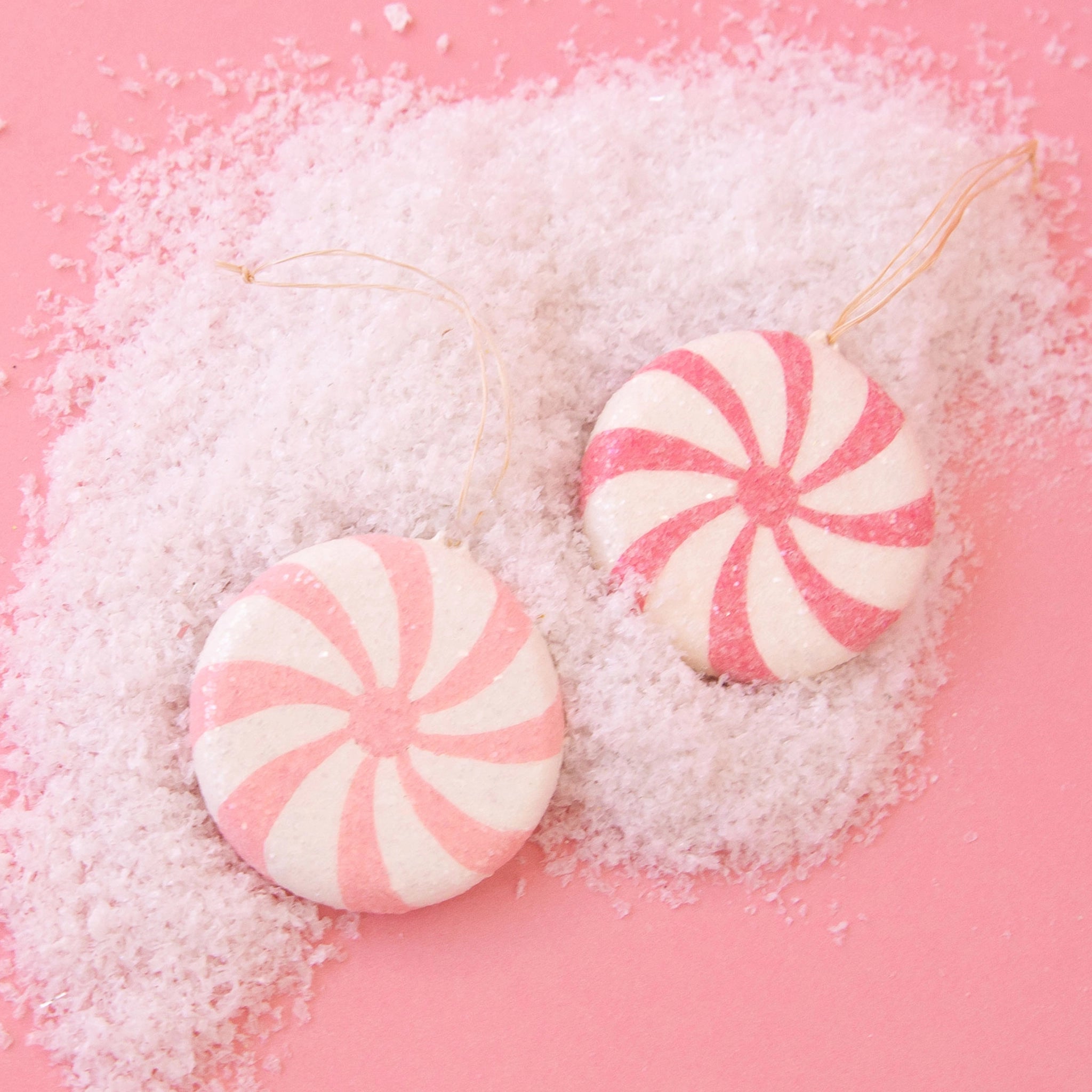 On a pink snowy background is two peppermint shaped ornaments. 