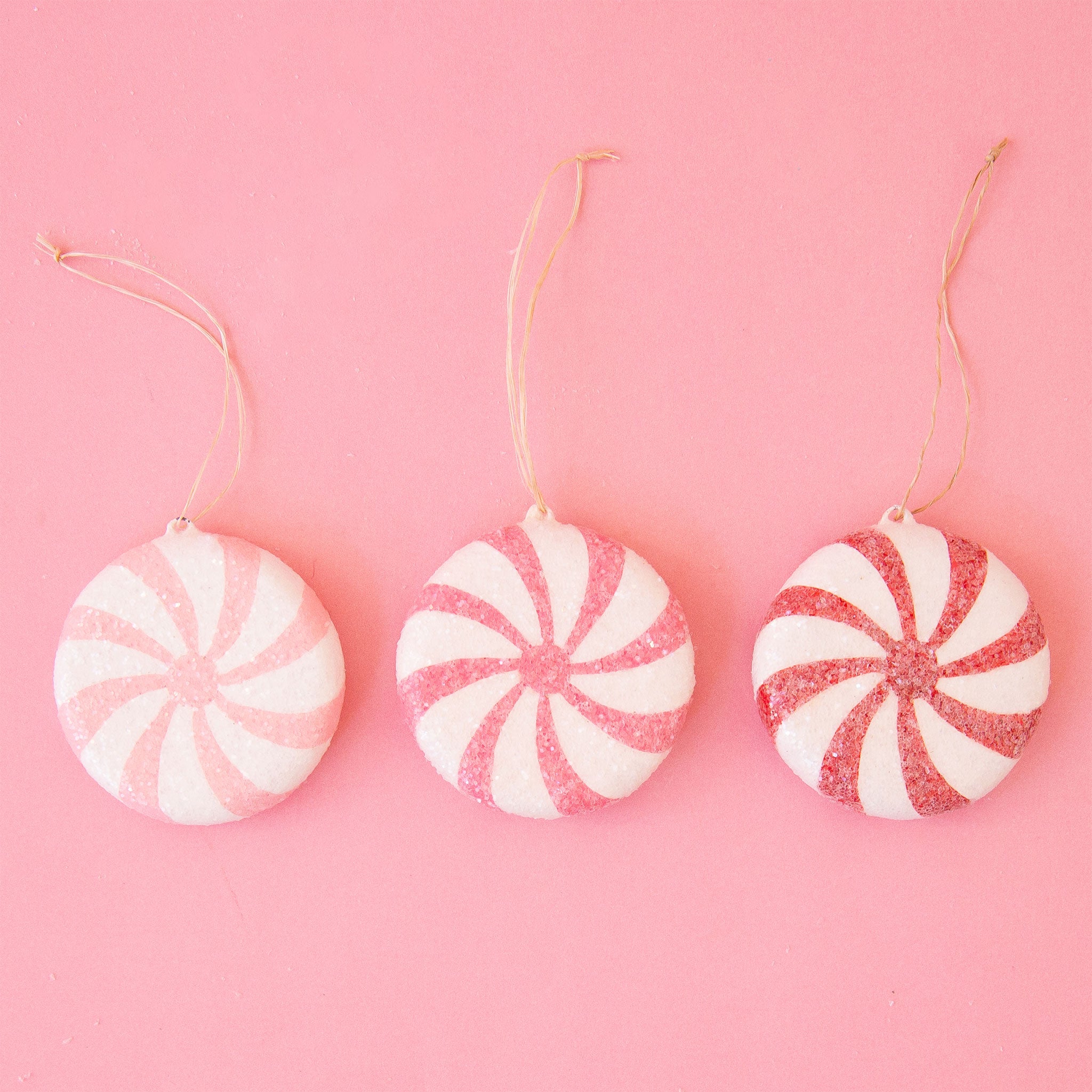 On a pink snowy background is three peppermint shaped ornaments.