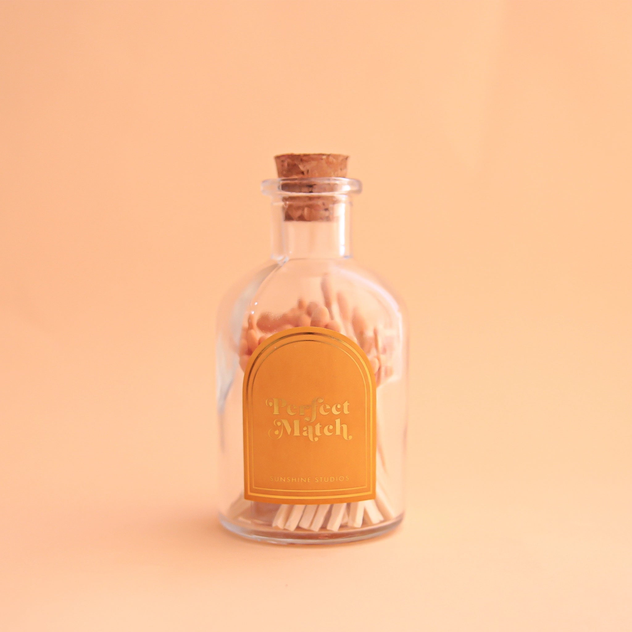 On a peachy background is a clear jar of wooden matches with a gold arched label on the front that reads, "Perfect Match". 