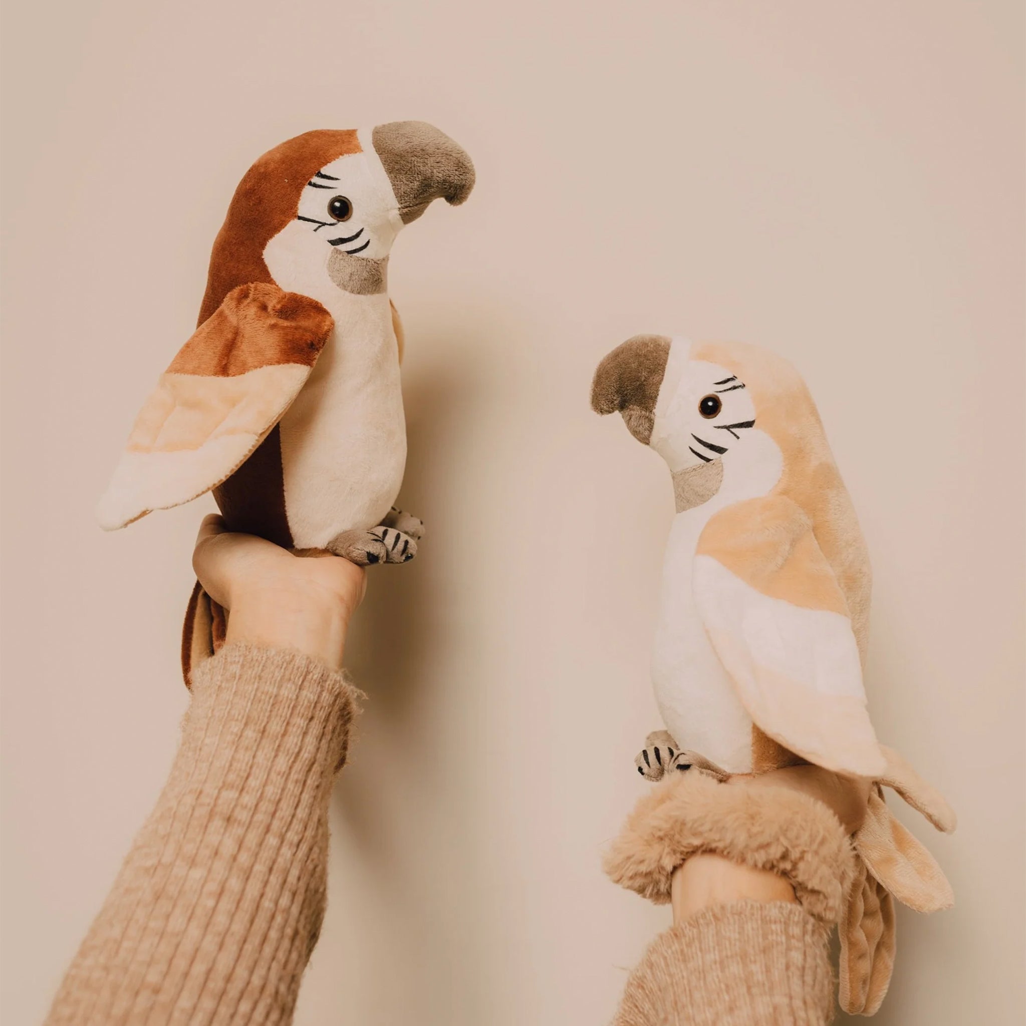 Two plush parrot toys held by hands against a beige background