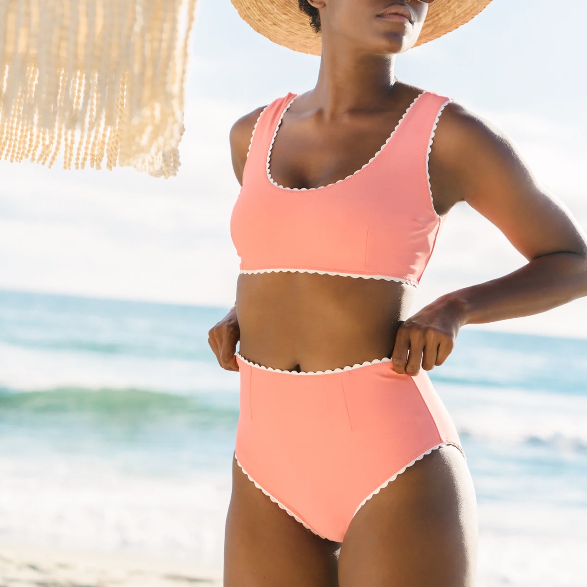 A model wearing a 1960's inspired swim suit with a wavy edge detail and high waist bottoms. The color of the swim suit is a coral pink hue.