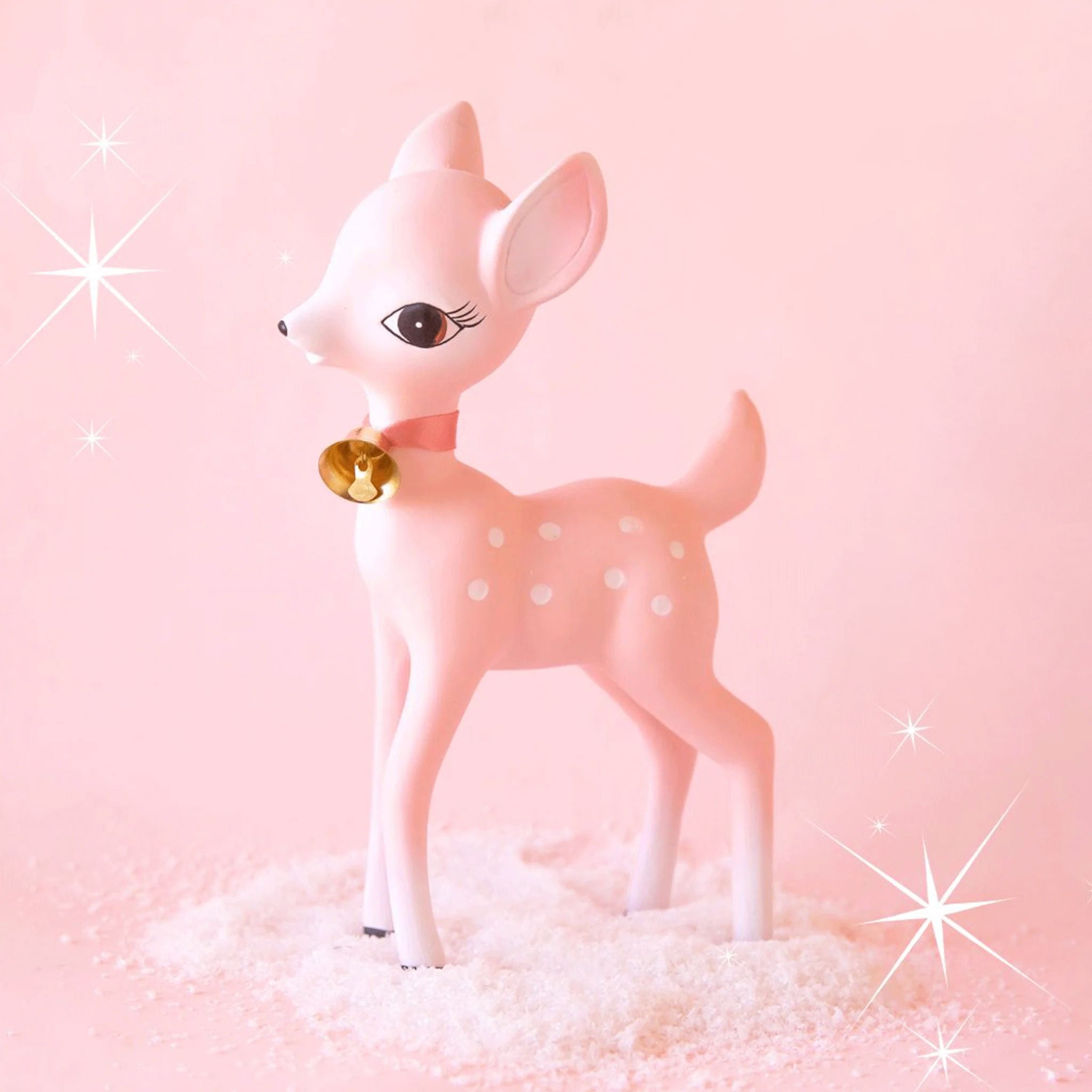 On a pink background is a retro deer shaped figurine in a pink shade with a gold bell around its neck and white spot details. 