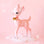 On a pink background is a retro deer shaped figurine in a pink shade with a gold bell around its neck and white spot details. 
