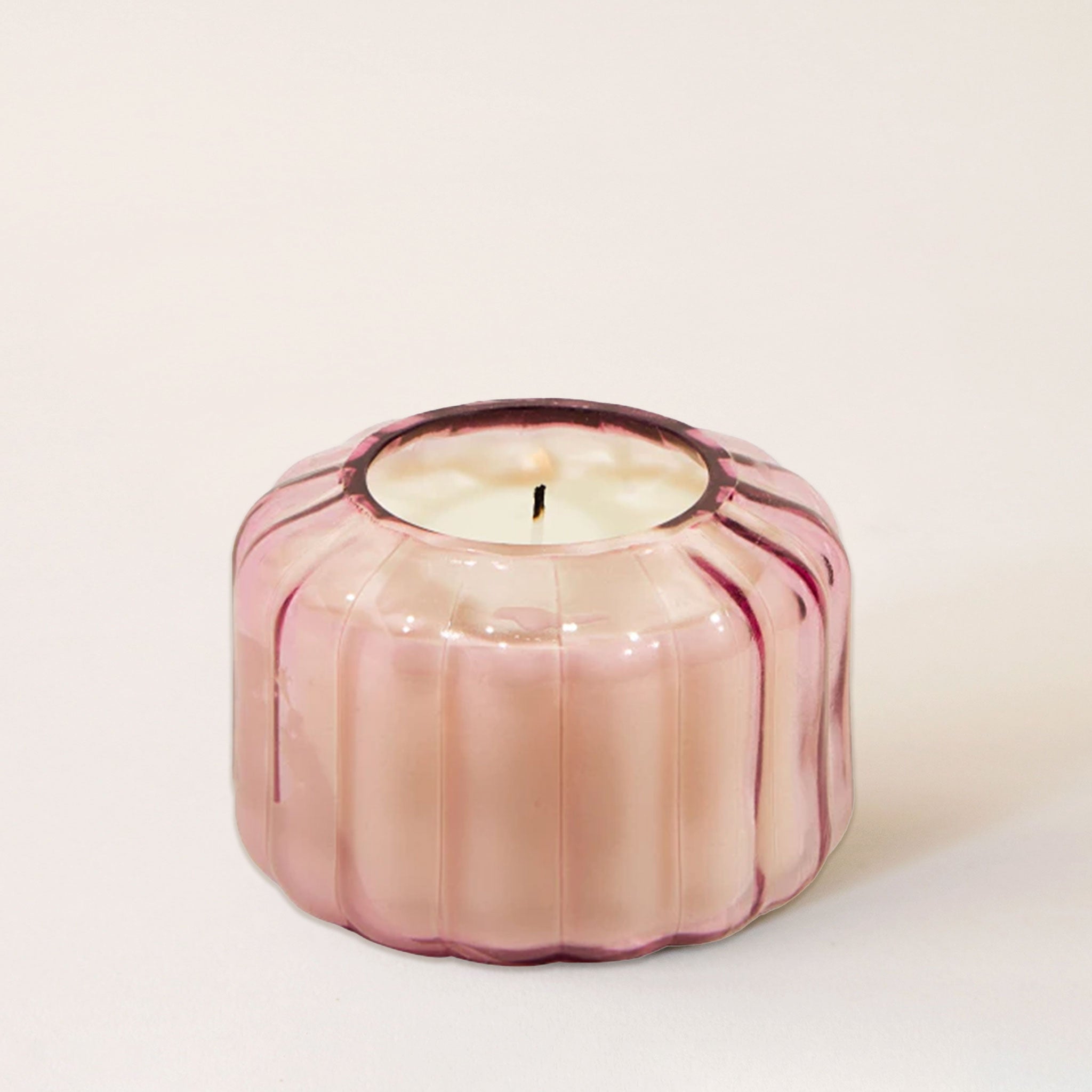 On a white background is a pink glass candle with a ribbed texture and a single wick.