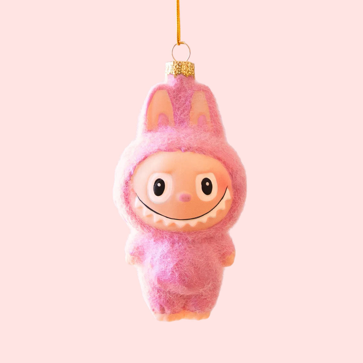 A small pink fuzzy monster ornament with bunny-like ears and a wide toothy smile hangs from a gold string against a soft lavender-pink background.