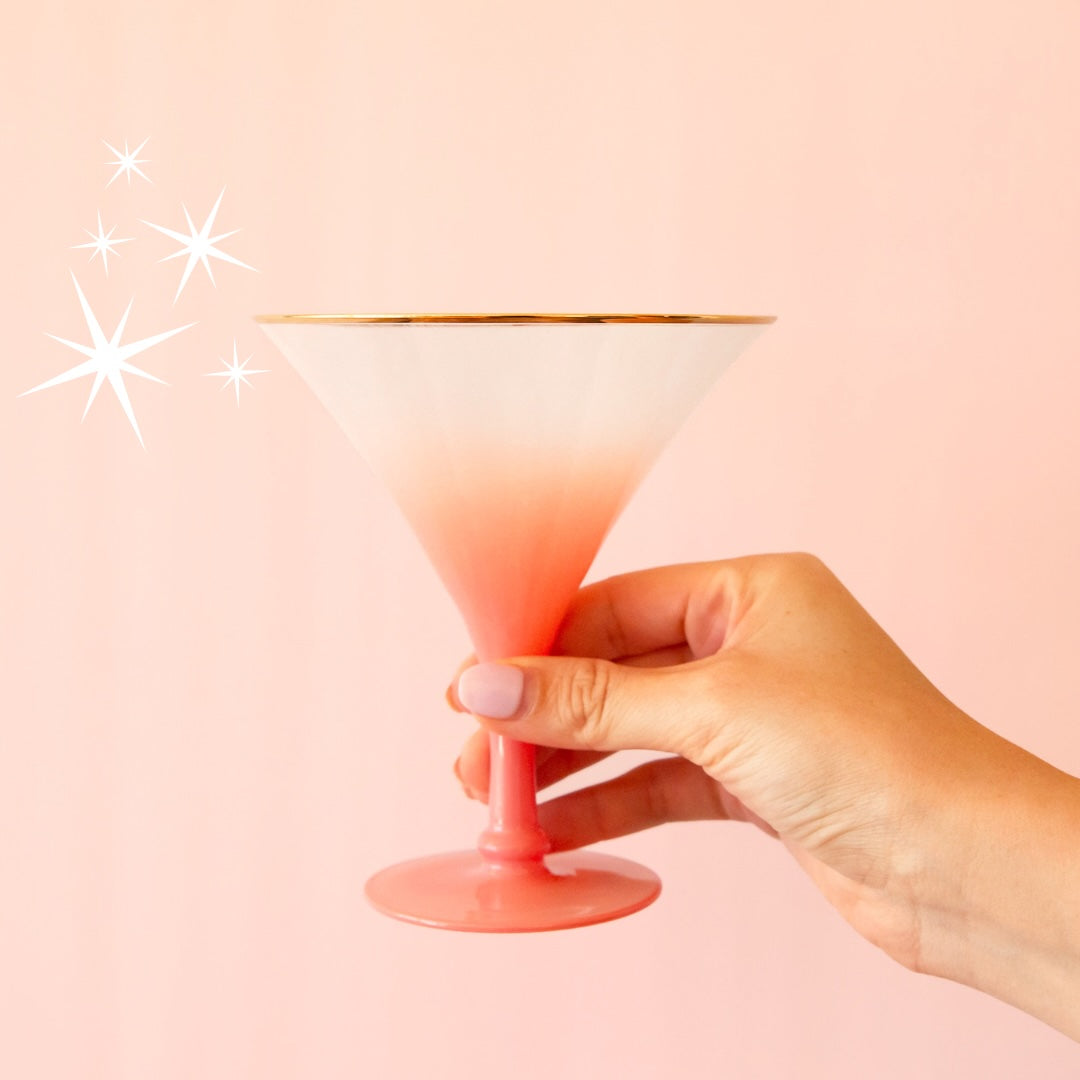 Hand holding a pink to white ombre cocktail glass with a shiny gold rim. 