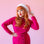 On a pink background is a model wearing pink santa hat with white faux fur detailing.