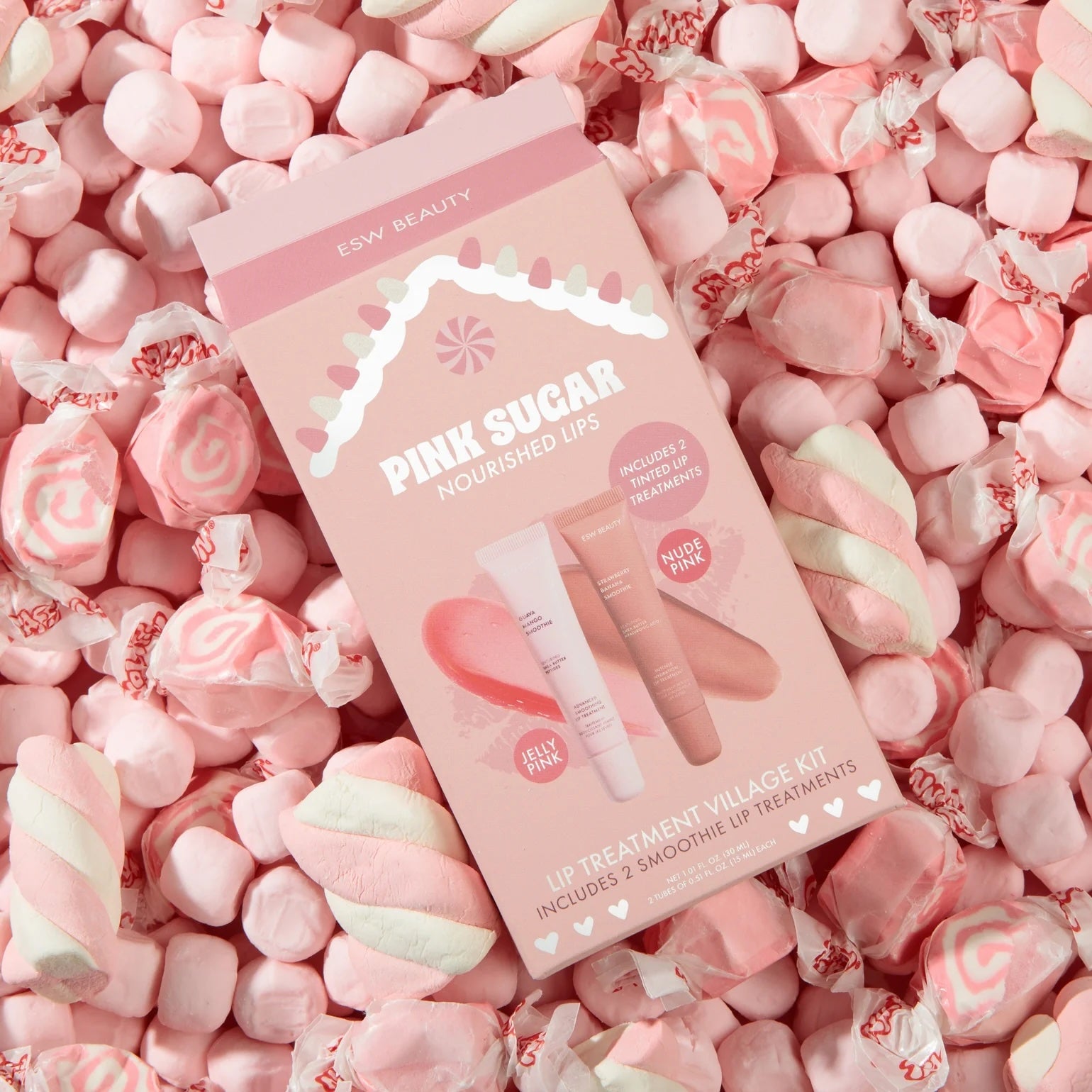 ESW Beauty ‘Pink Sugar Nourished Lips’ lip treatment kit box resting on a pile of pink marshmallows and wrapped candies in a pastel, candy-themed flat lay.