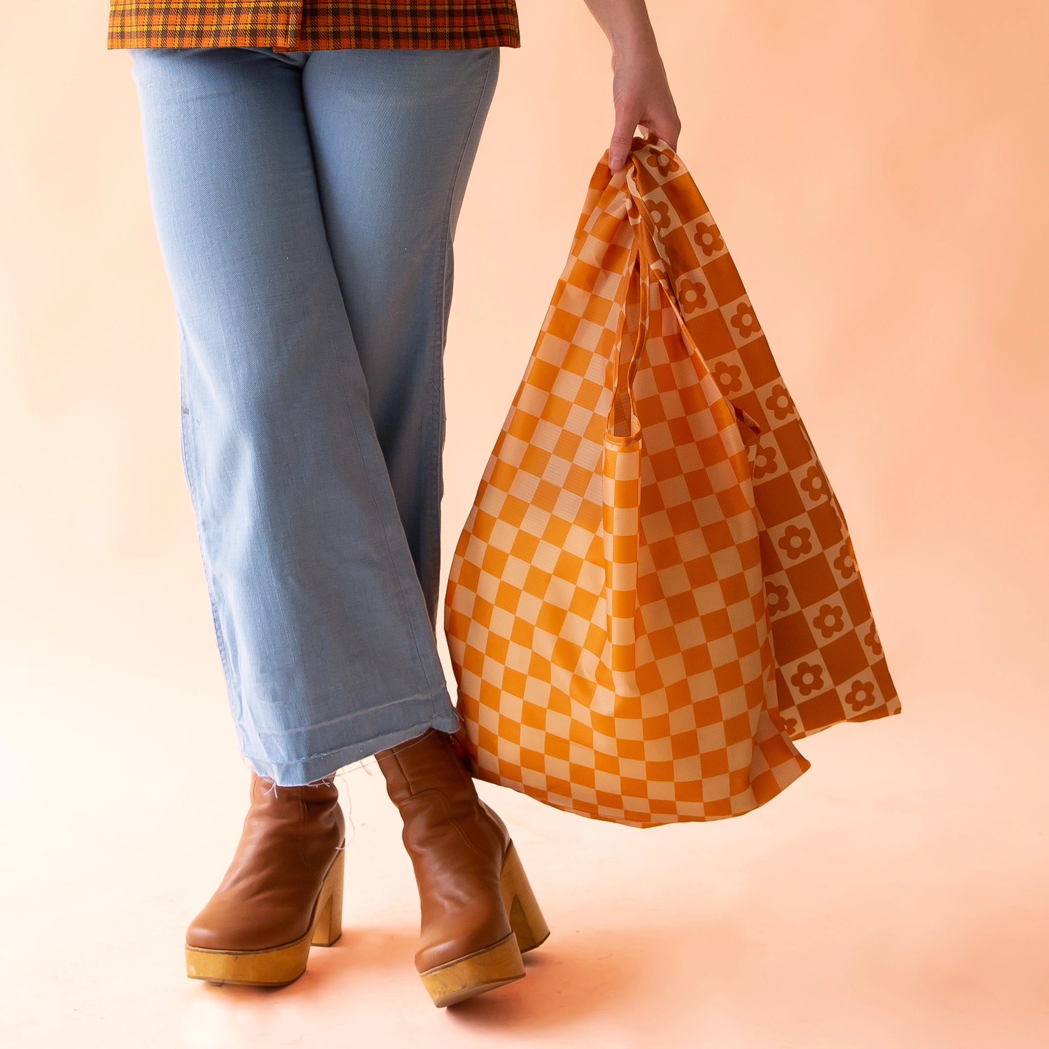On a light peach background is a model holding two reusable nylon bags. One is a dark and light orange checkered print and the other is a burnt orange and light orange checker print with flowers in the squares.