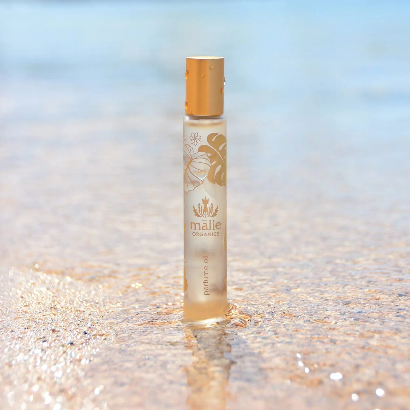 A gold rollerball perfume that reads "Maile organics perfume oil".  The roller sits in sand with ocean water flowing next to it.
