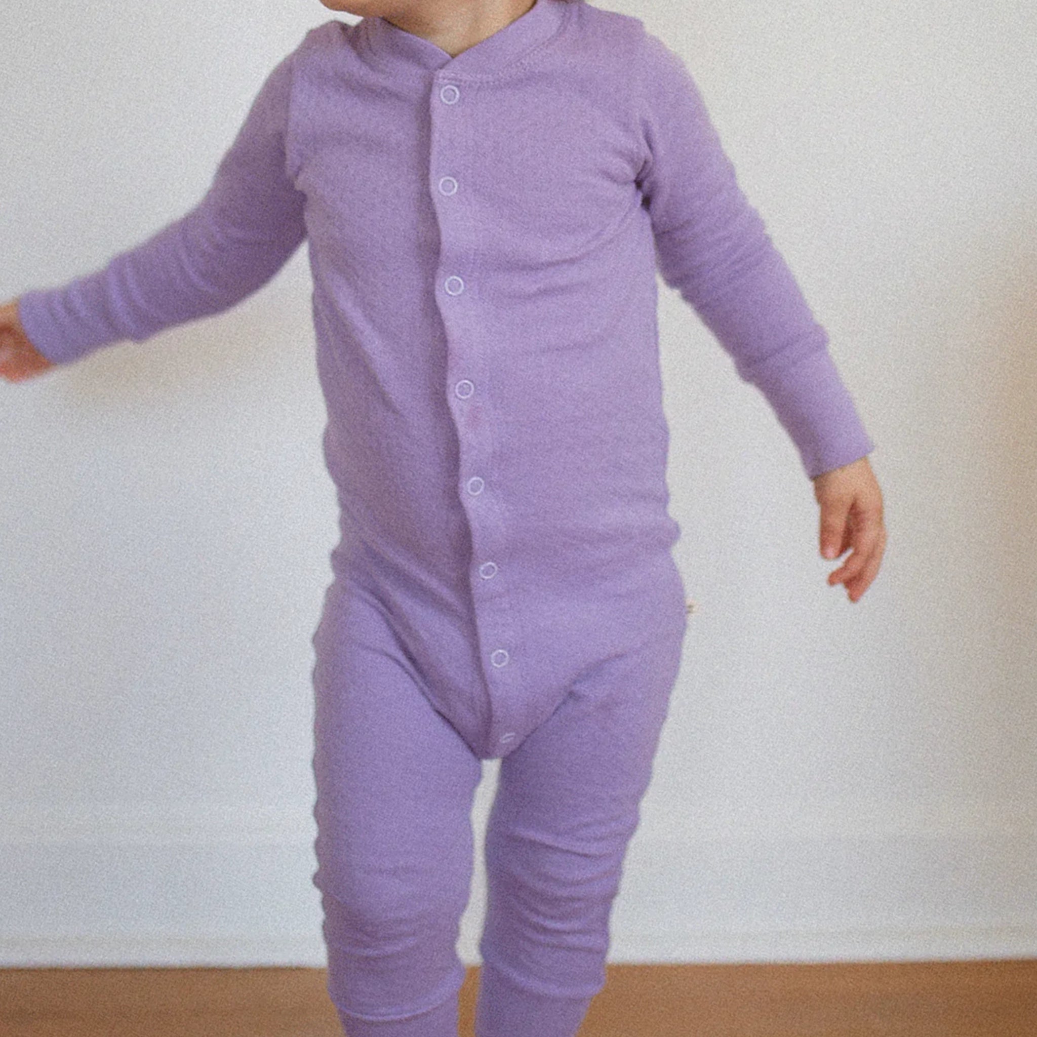 On a white background is a baby / toddler model wearing the lavender one piece sleep set with buttons up the front. 