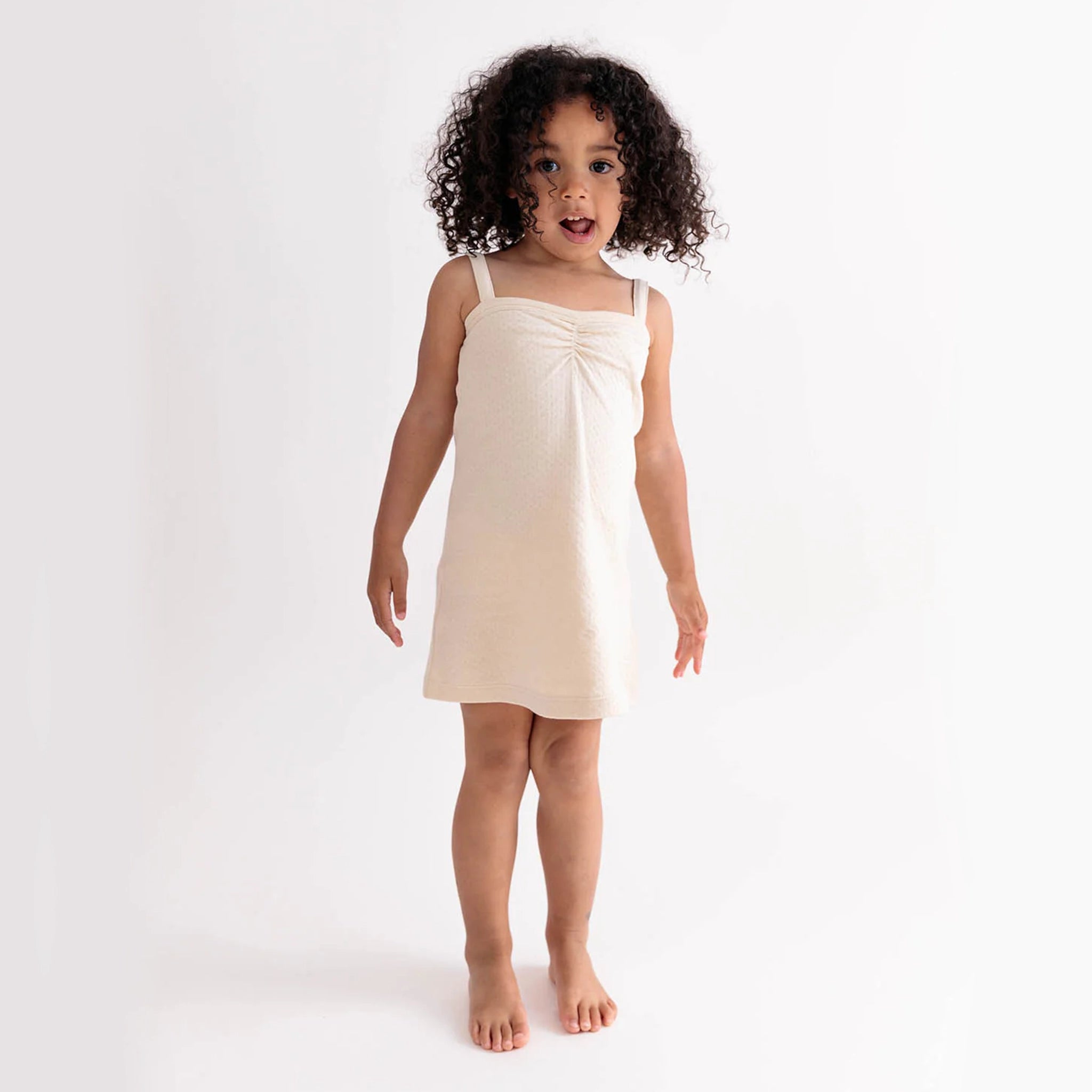 On a white background is a child model wearing an ivory dress. 