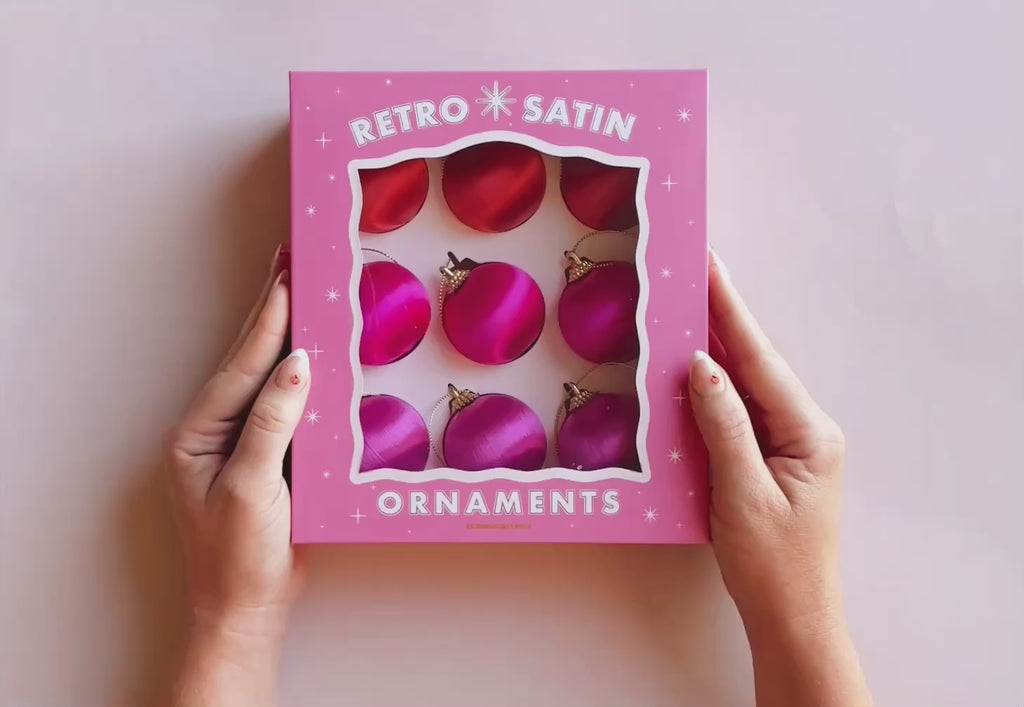 Hands opening a pink box that reads "retro satin ornaments".  Inside the box are 9 round pink and orange hue satin ball ornaments.