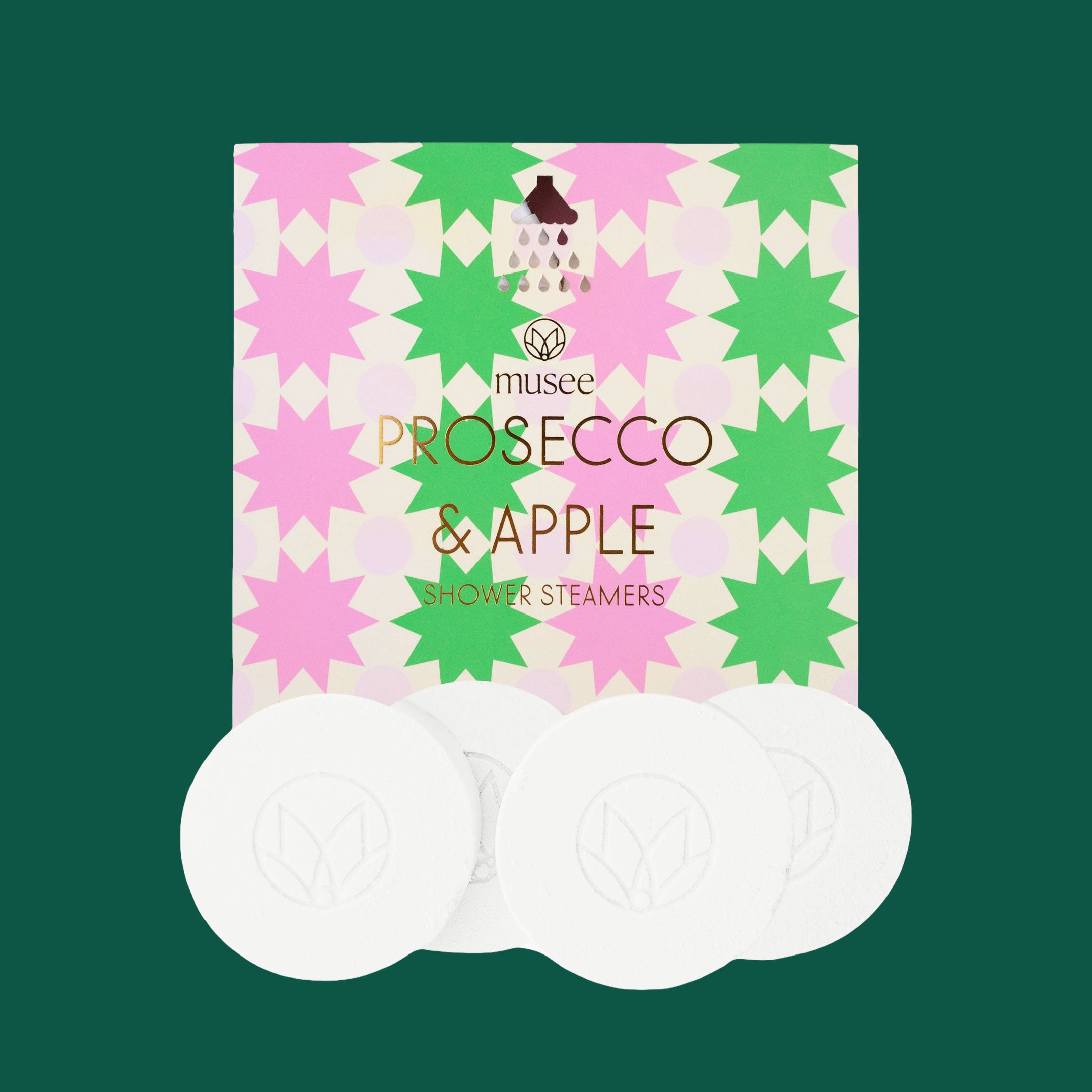 A colorful box of “Musee Prosecco & Apple Shower Steamers” displayed against a dark green background. The packaging features a vibrant pattern of pink and green starburst shapes with gold lettering. Four round white shower steamers with an embossed leaf logo are arranged in front of the box.