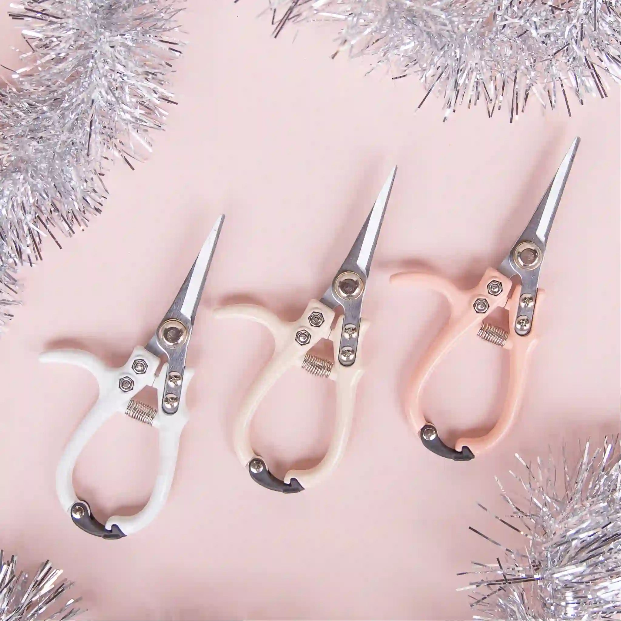 All three color ways of the pruning shears photographed next to each other and staged in the middle of silver tinsel. There is a white, a light pink and a neutral tan.