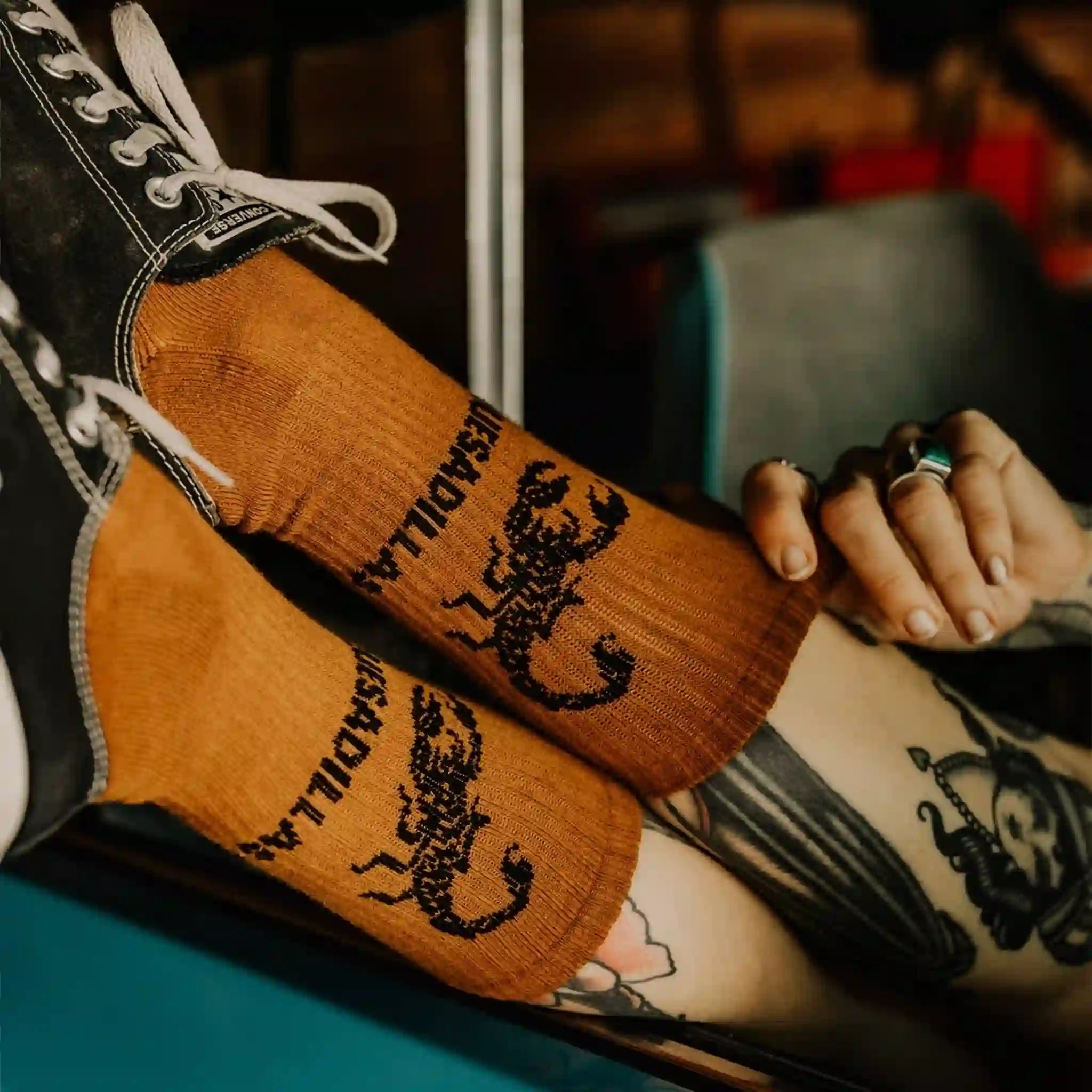 A model wearing a pair of burnt orange crew socks with a black scorpion graphic and text that reads, "Quesadilla".