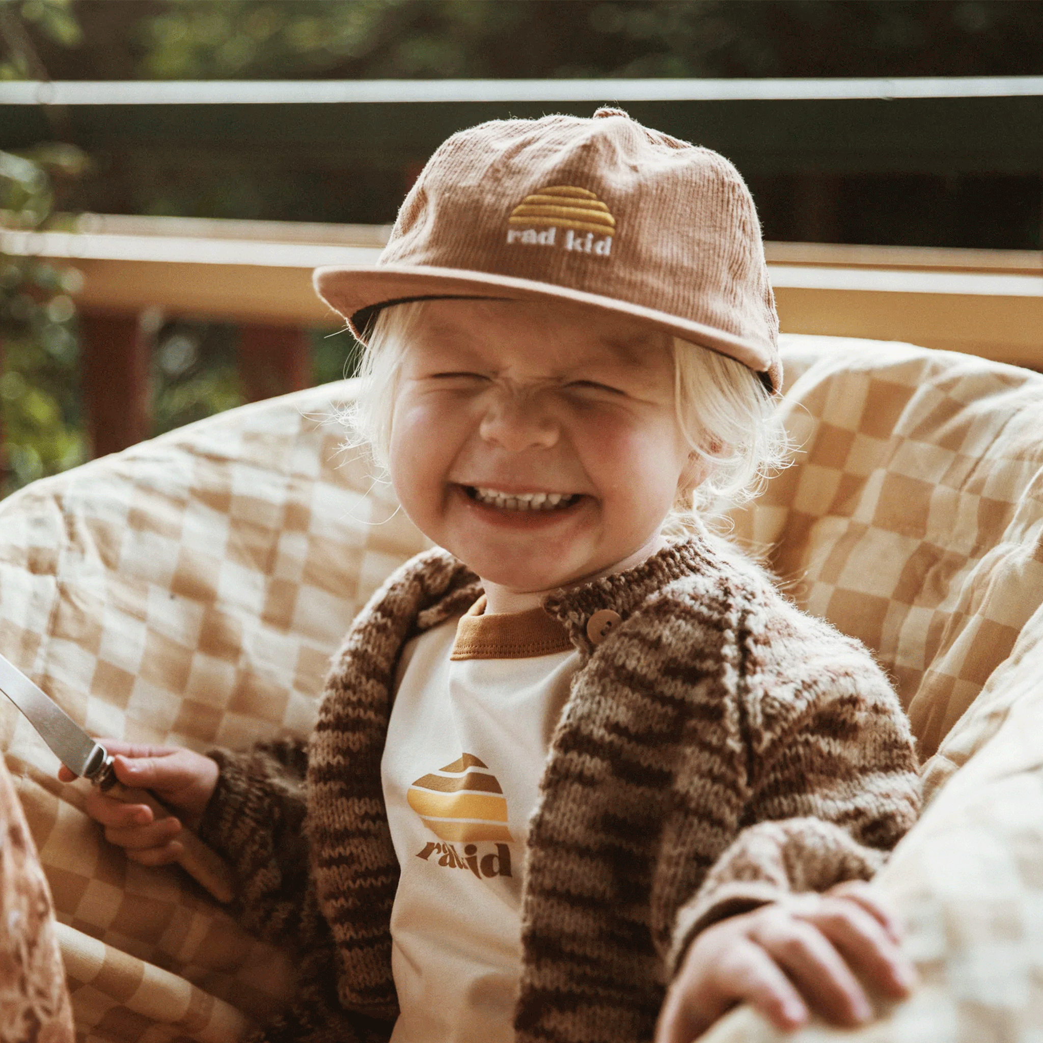 A children&#39;s model wearing a tan corduroy hat with a yellow sun logo and white text below it that reads, &quot;rad kid&quot;.