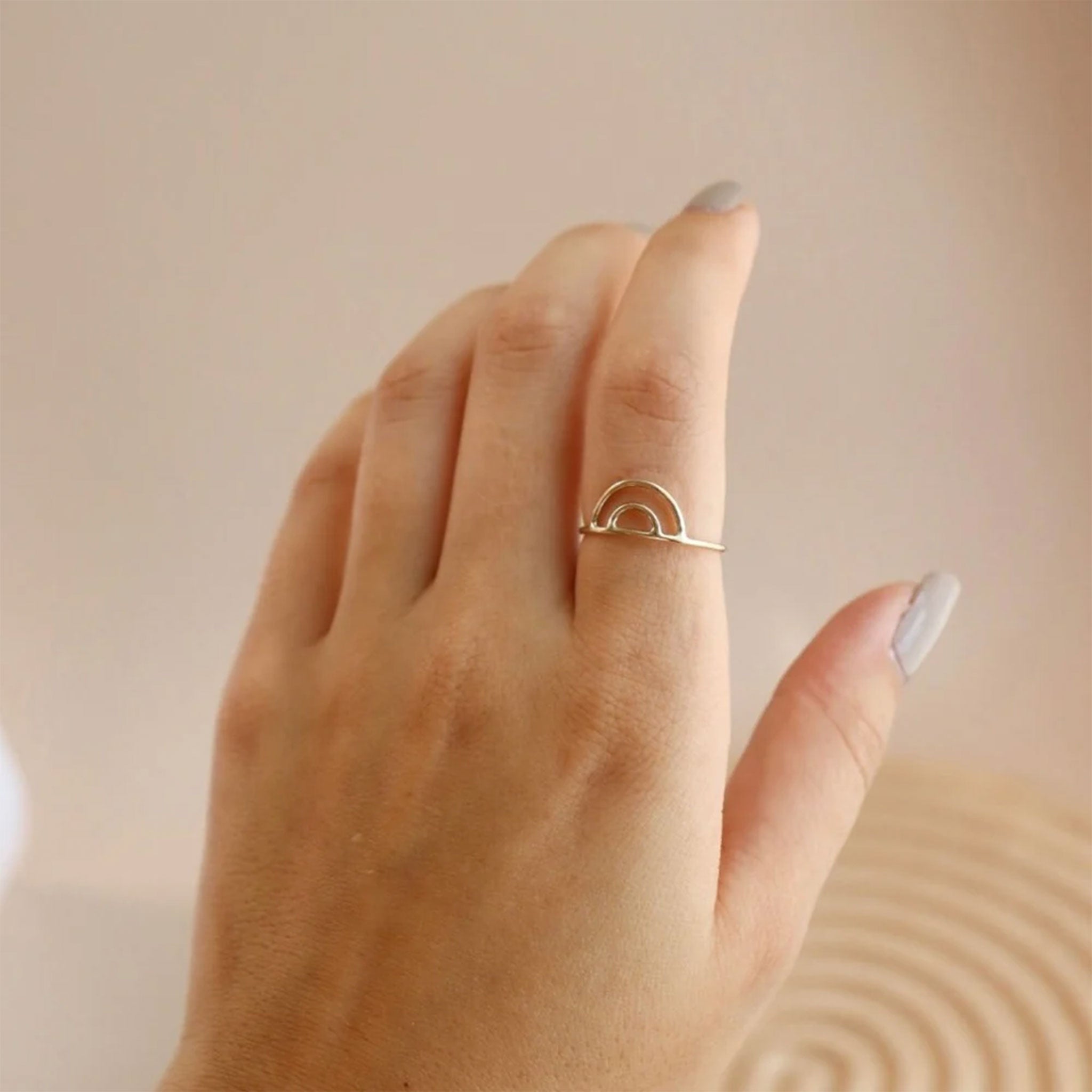 On a tan background is a model&#39;s hand with a dainty gold rainbow shaped ring. 