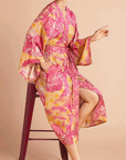 On a tan background is a model wearing a pink and yellow/orange robe with a tropical and floral print.