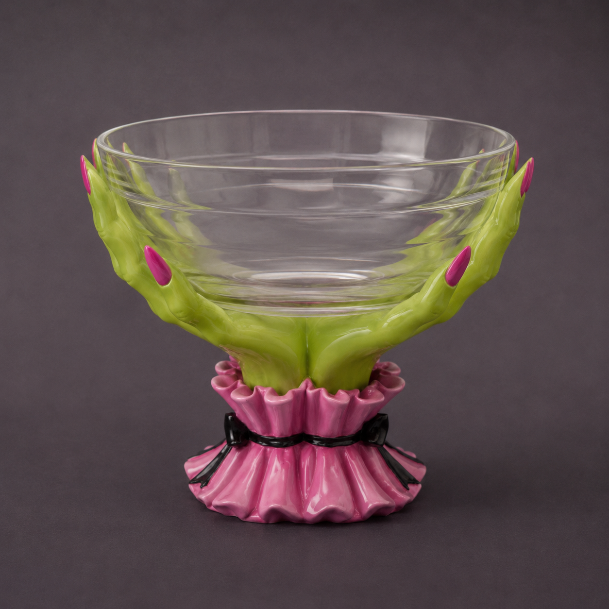 A decorative clear glass bowl with sculpted green hands supporting it, accented with pink fingernails and a pink ruffled base tied with a black ribbon, shown on a dark gray background.