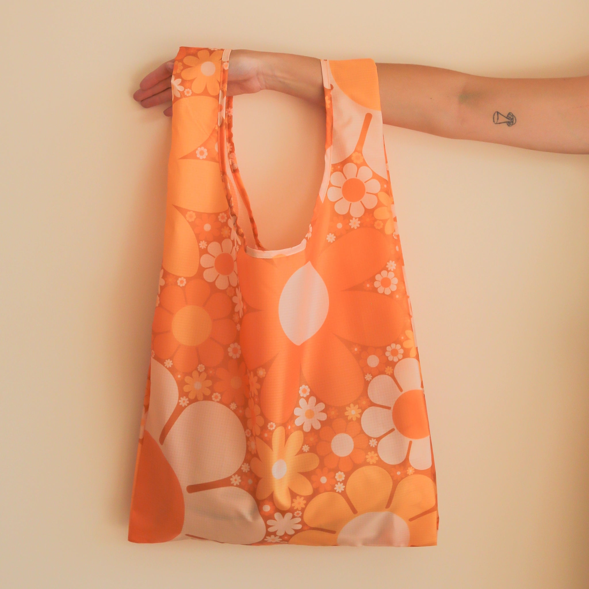 On a tan background is a model's arm holding a nylon reusable bag with an orange floral print. 