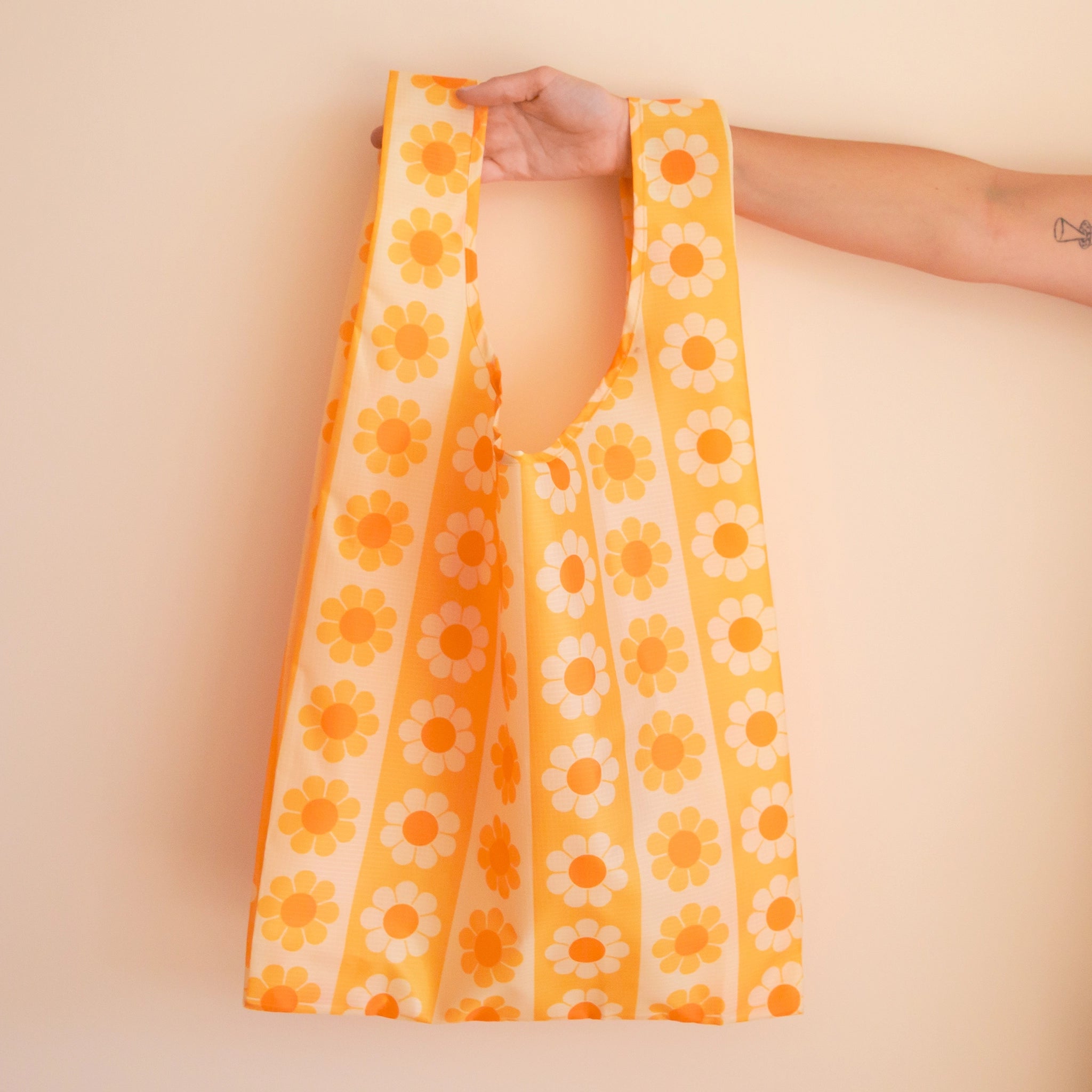 On a tan background if a model&#39;s hand holding out a reusable nylon bag with a yellow and orange daisy pattern.