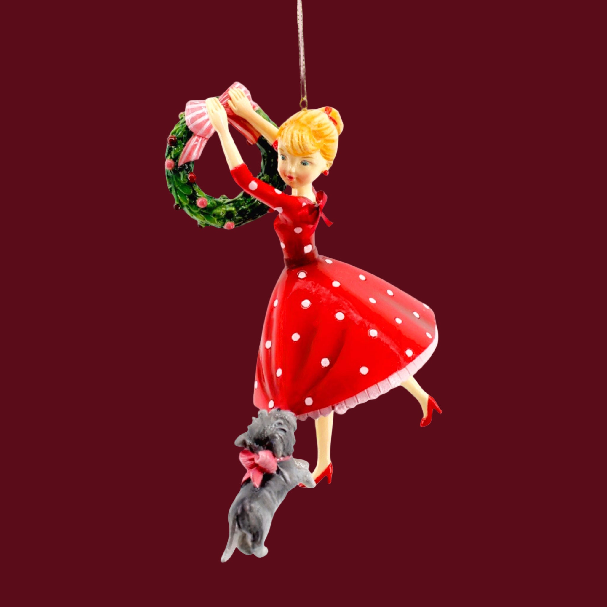 A retro looking girl wearing a big red dress with polka dots.  A gray dog tugs at her dress while she hangs a Christmas wreath.