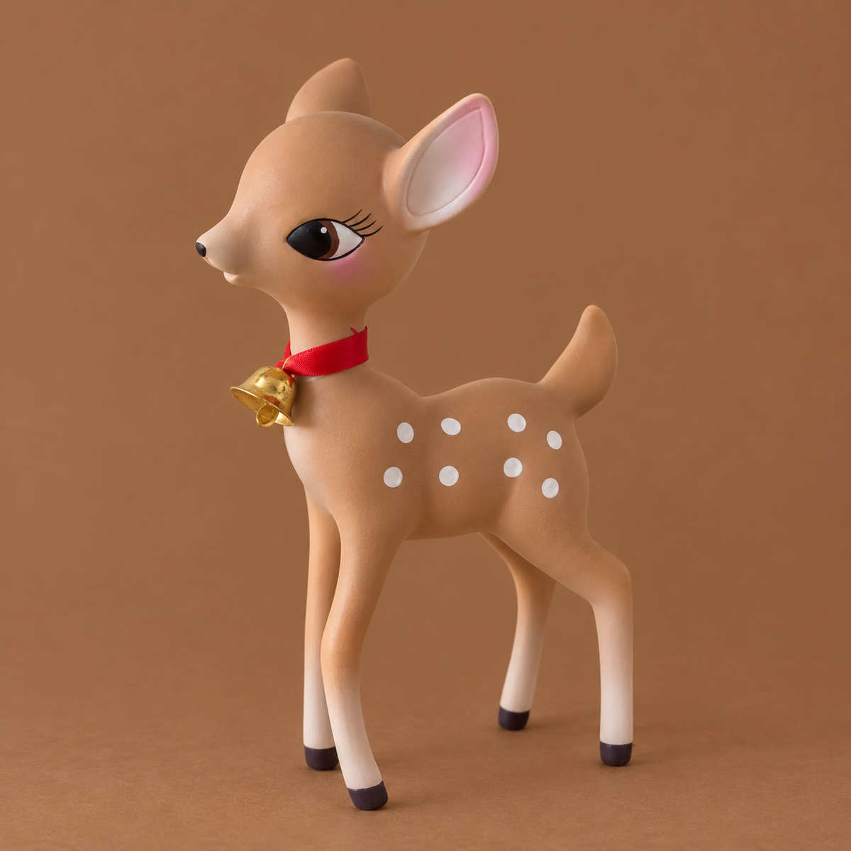 A stylized fawn figurine with a tan body, white spots, pink inner ears, a red collar, and a small gold bell, photographed against a warm brown studio background.