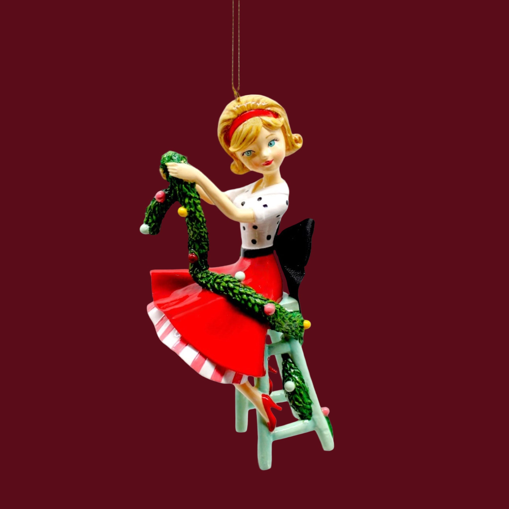 A resin ornament of a retro style girl wearing a red skirt and polka dot top.  She is holding green Christmas decorating garland and sitting on a mint stool.
