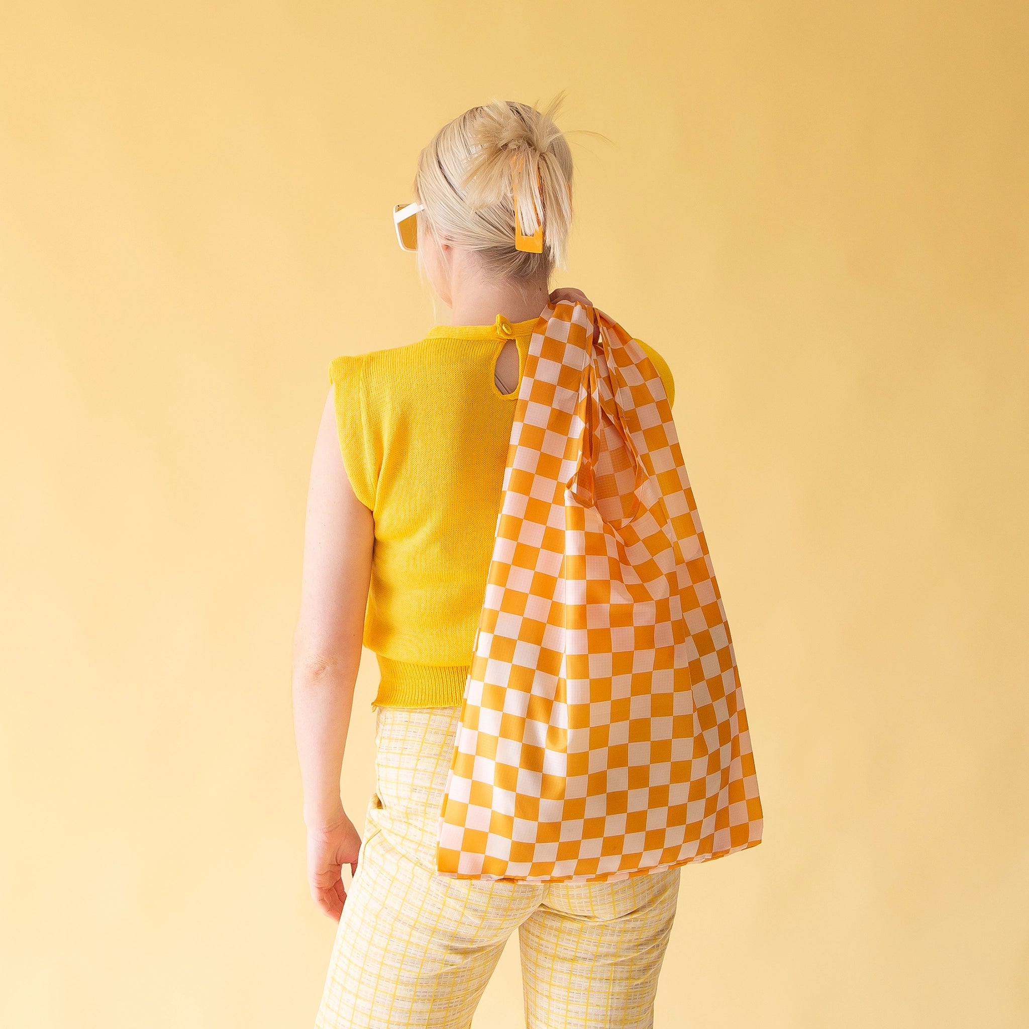On a light yellow background is a model holding the light orange and ivory checkered printed nylon bag over their shoulder.