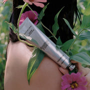 Close-up of a silver tube of FAMT Estate Rich Cream (Jasmine Damask Rose) resting against a person’s shoulder, surrounded by green leaves and pink flowers in a garden setting.
