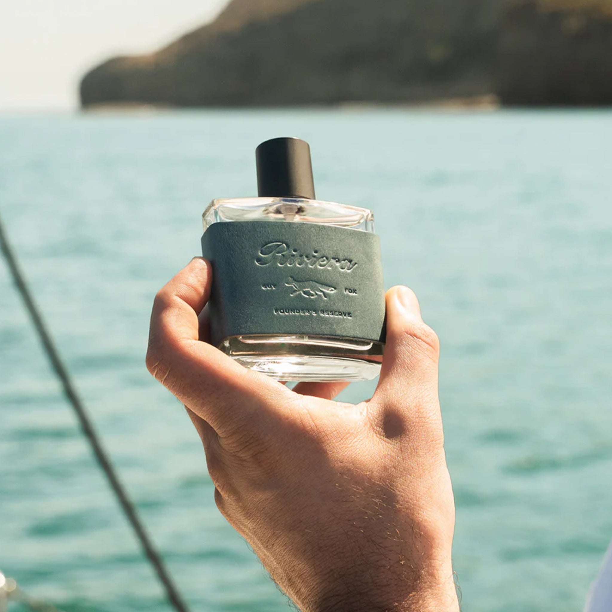 Hand holding a perfume bottle with a scenic background of water and mountains.