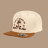 A cream colored kids snapback with brown bill and brown embroidery that reads, 'Surf and Skate Co BNKY SURF' and a graphic of a palm tree'. 