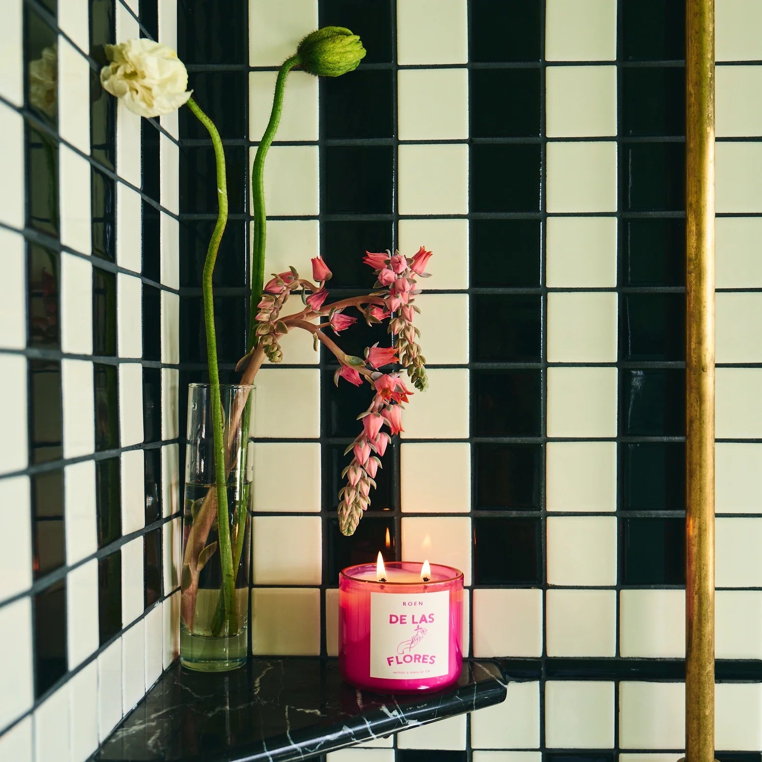 A pink glass candle that reads "De Las Flores" on a black and white til background with flowers next to it.