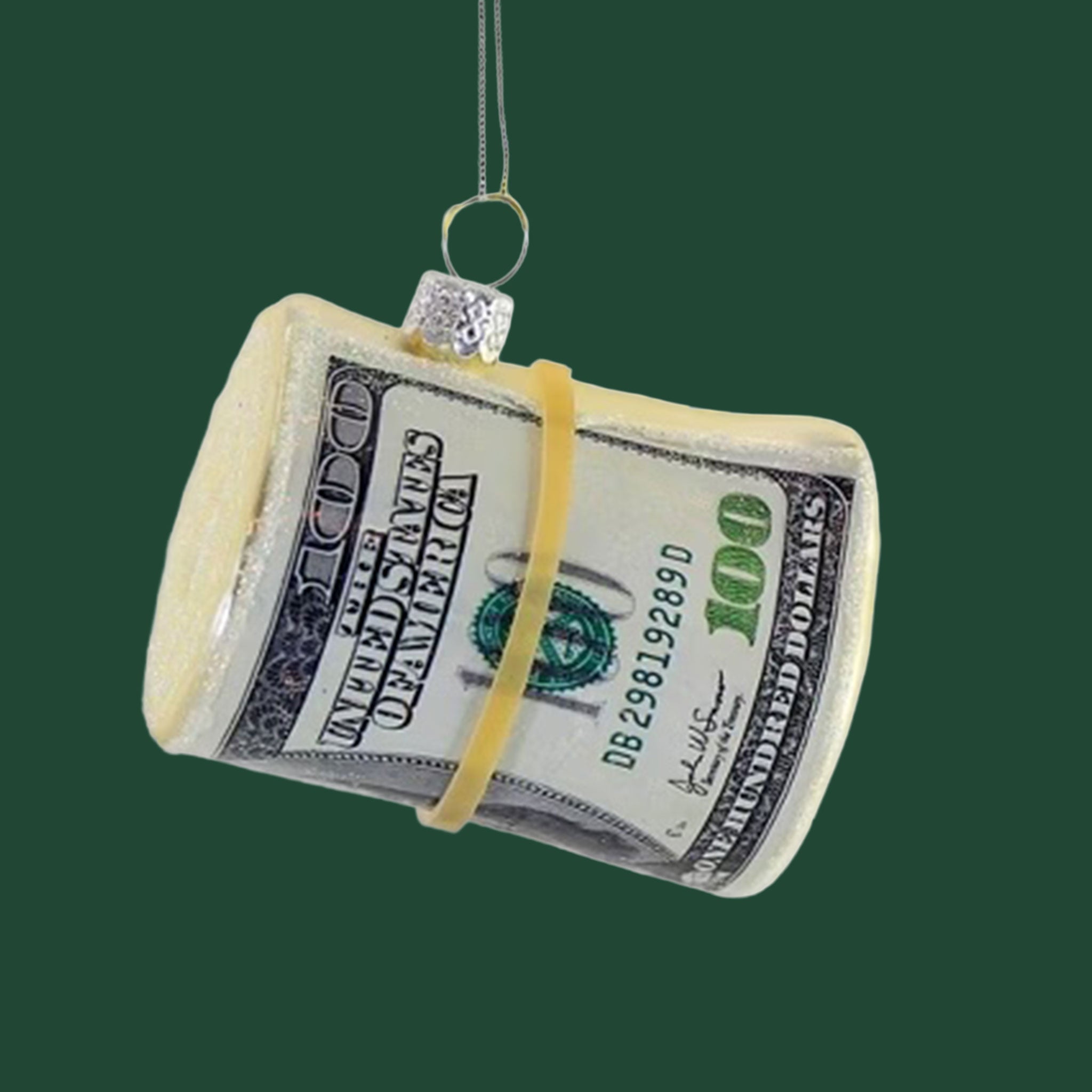 A roll of hundred dollar bills with a rubber band around them sits on a dark green background.  The roll of cash is fake and has a silver string tied to it, the roll of cash is a Christmas ornament.