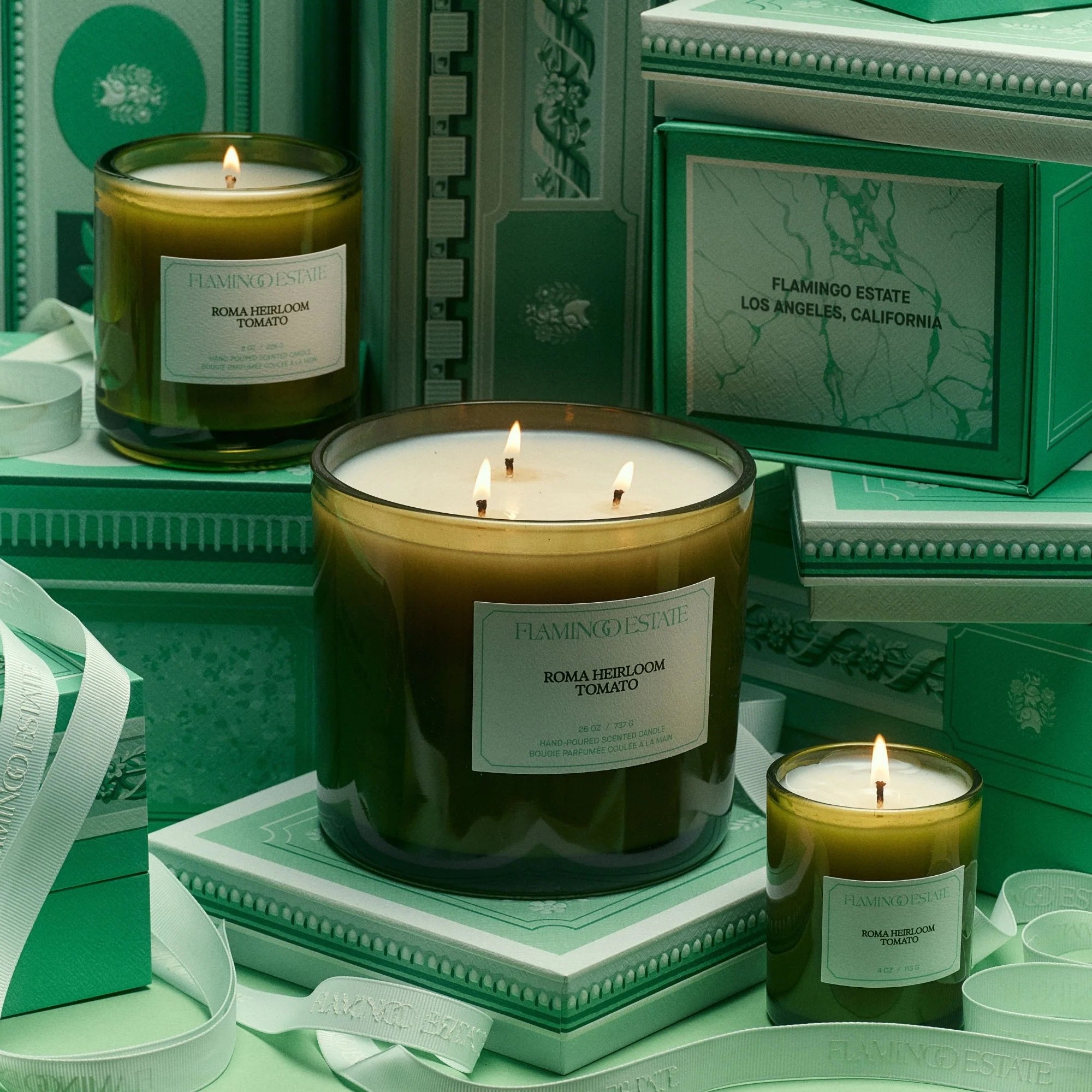 Three green glass candles labeled ‘Flamingo Estate – Roma Heirloom Tomato’ burn among stacked green decorative boxes and ribboned packaging in a monochrome green scene.
