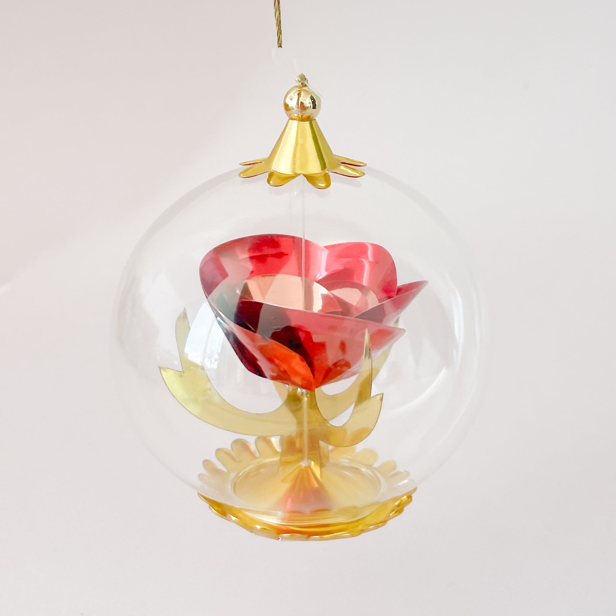 A clear round glass ornament hangs on a white background, containing a stylized red rose with gold leaves and a gold base, topped with a small gold cap.