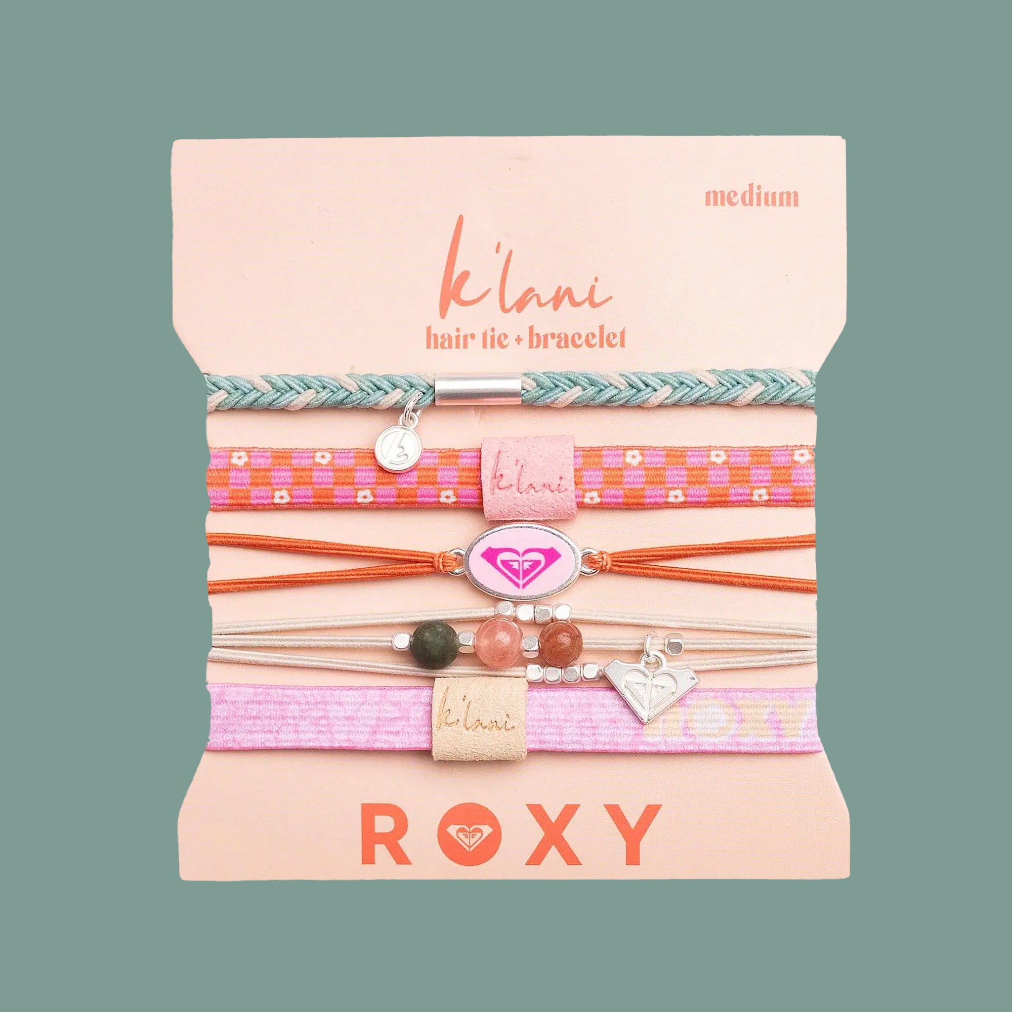 A set of five Roxy K'lani hair tie bracelets in various colorful woven and cord patterns, presented on their pink cardboard packaging against a muted green background.