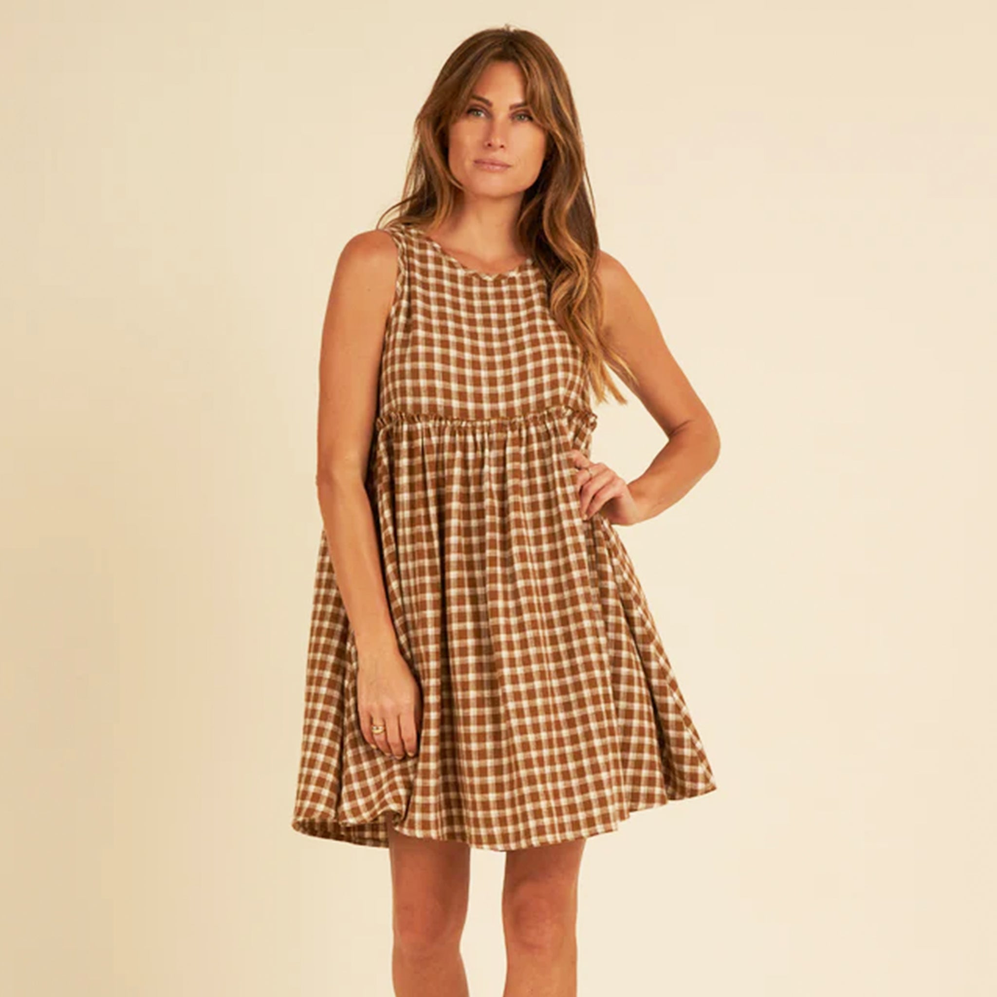 On a tan background is a model wearing a brown and tan plaid babydoll dress with a tie detail in the back.