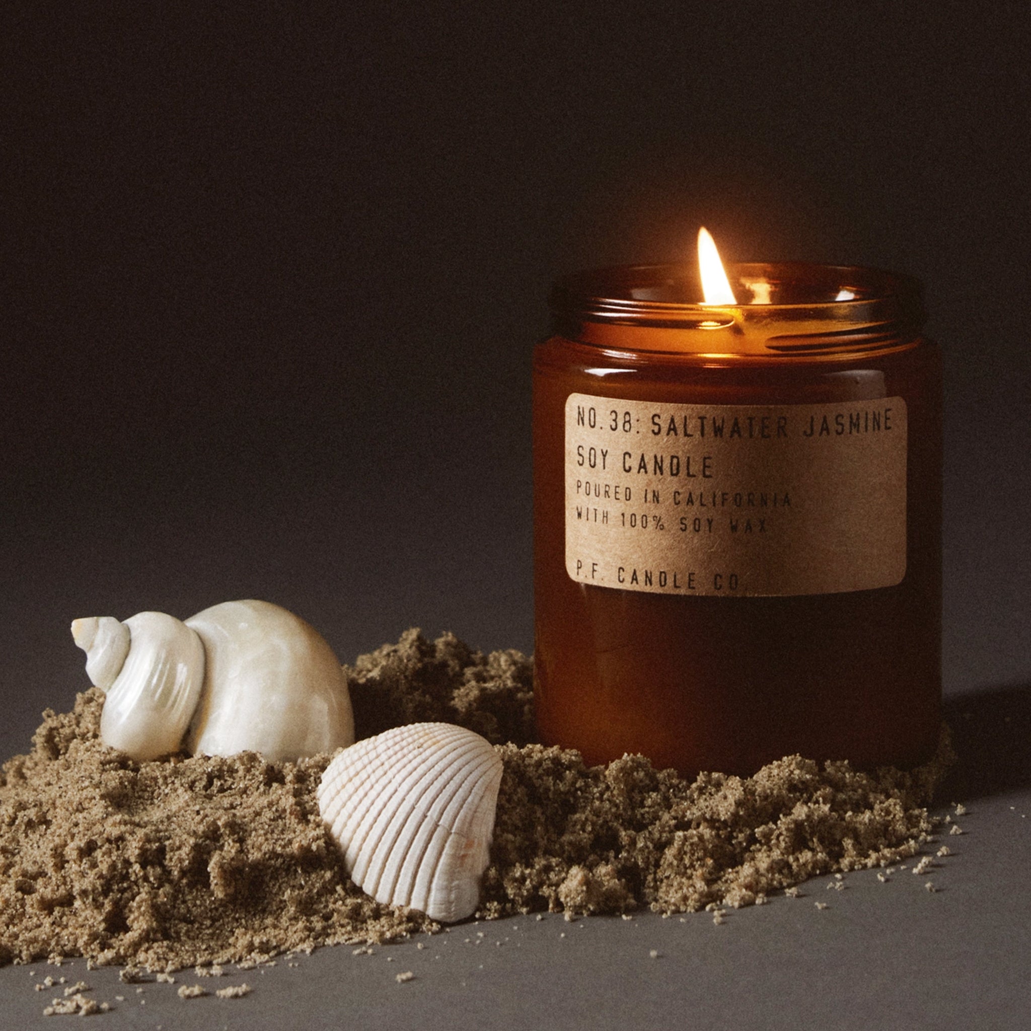 Candle in an amber jar with a label, surrounded by sand and shells on a dark background