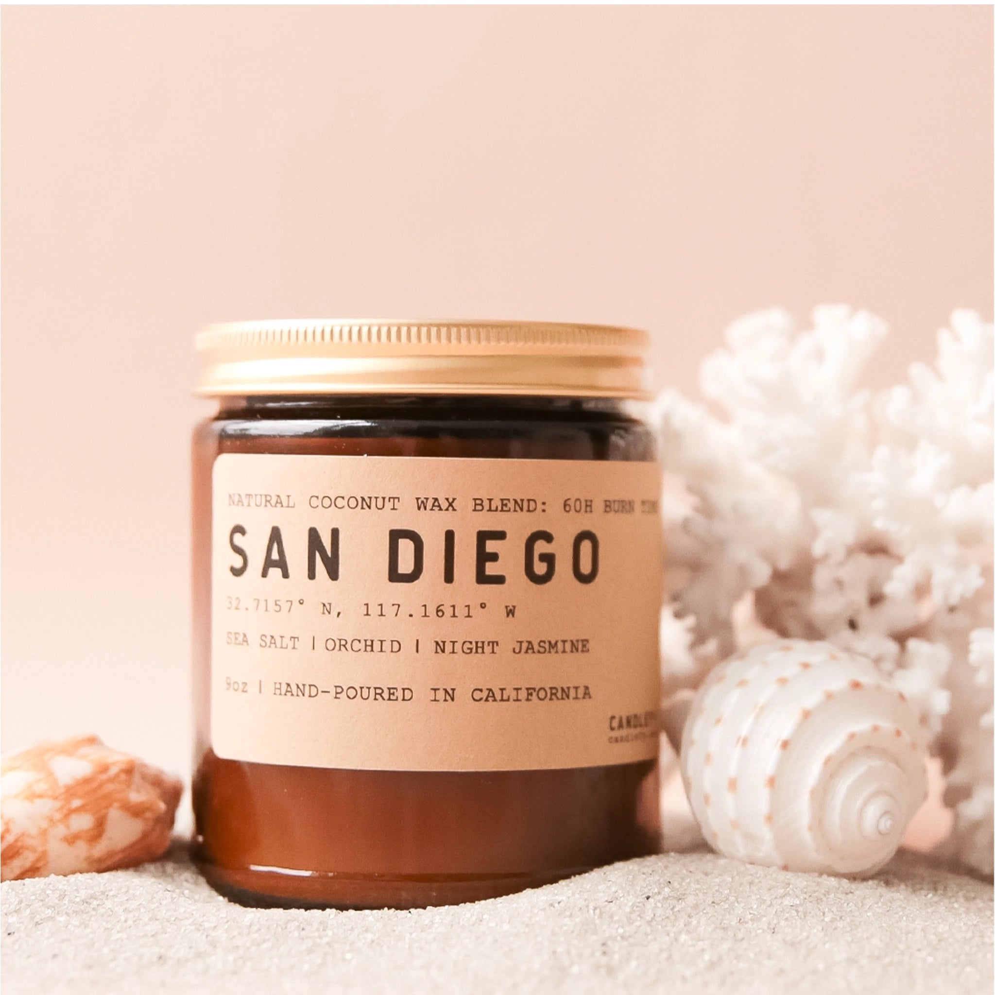 On a light pink background is a amber glass candle with a gold lid and a label that reads, "San Diego" and is staged next to beach inspired items like shells and sand. 
