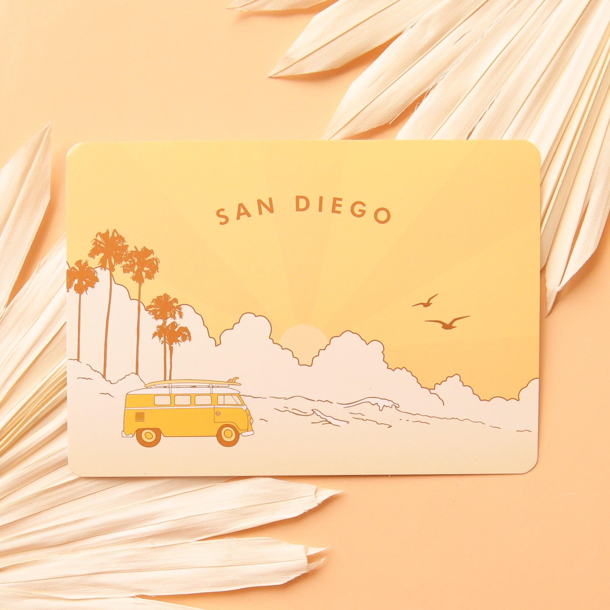 A yellow and cream postcard with a San Diego beach graphic design with palm trees and birds in the background along with a bright yellow VW bus that has a surf board on top as well as text curved along the top that reads, "San Diego".