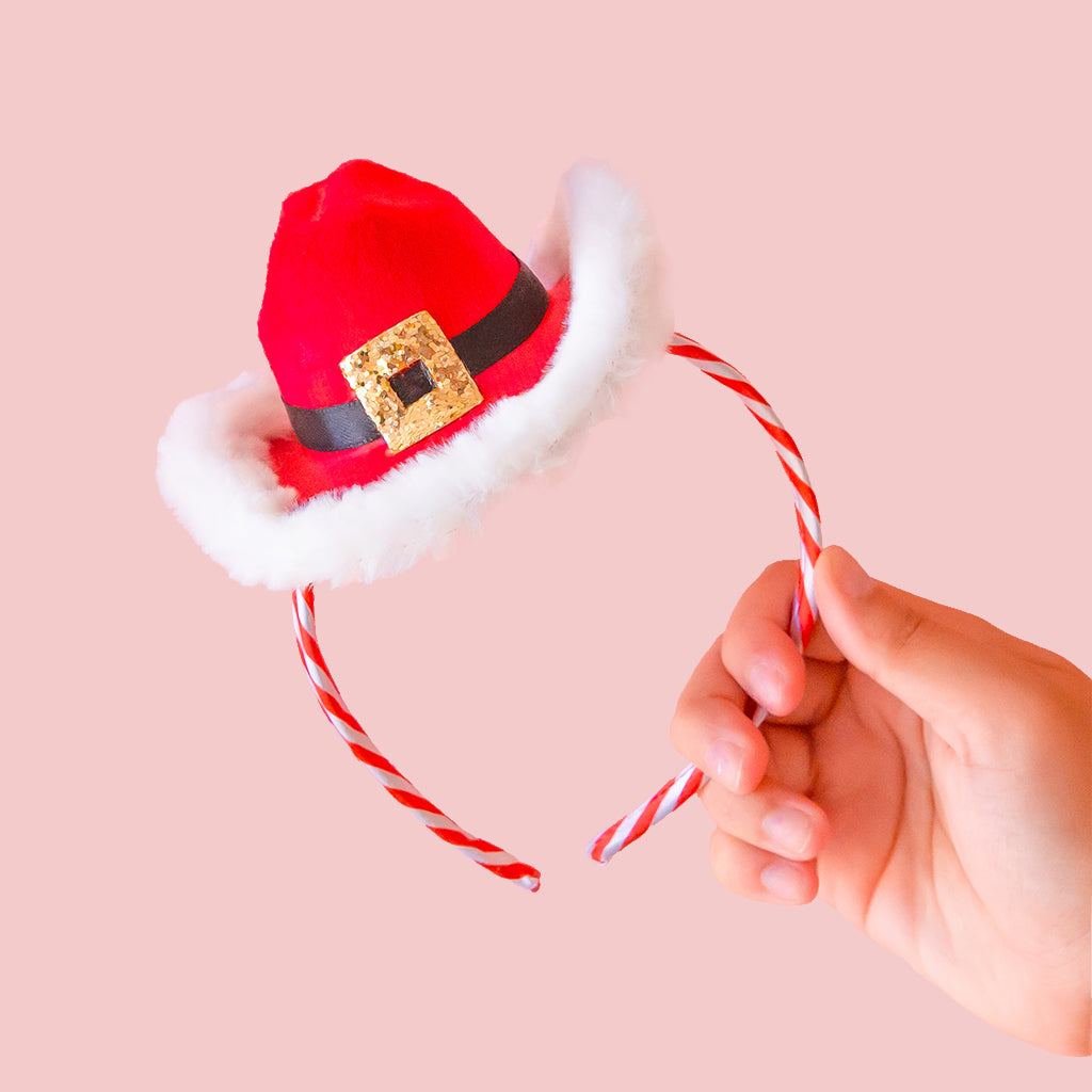 Hand holding a small Santa hat headband with candy canes detail on the headband against a pink background