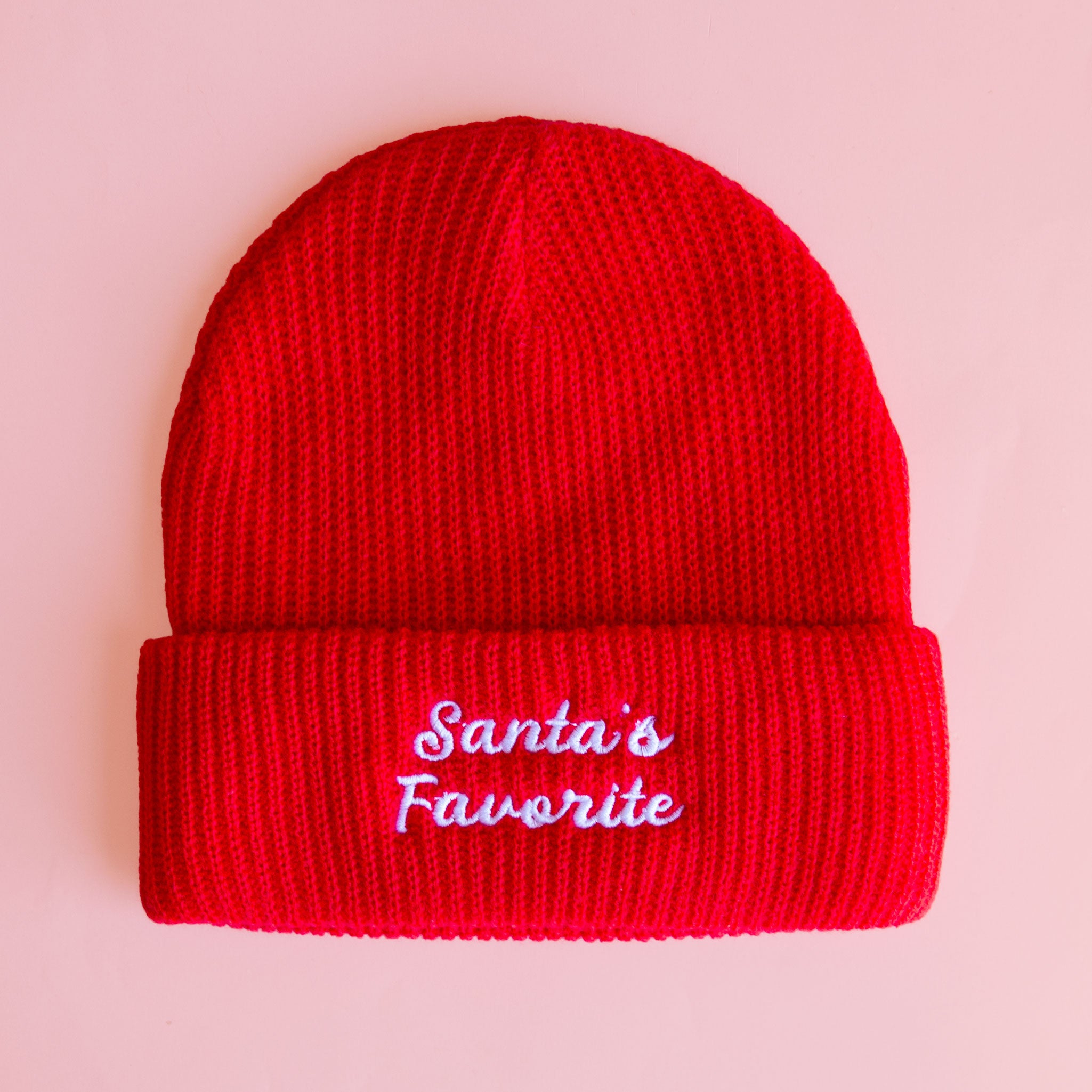 Product photo of a bright red knitted winter beanie, folded at the brim, with white cursive embroidery reading ‘Santa’s Favorite,’ centered against a pale pink backdrop
