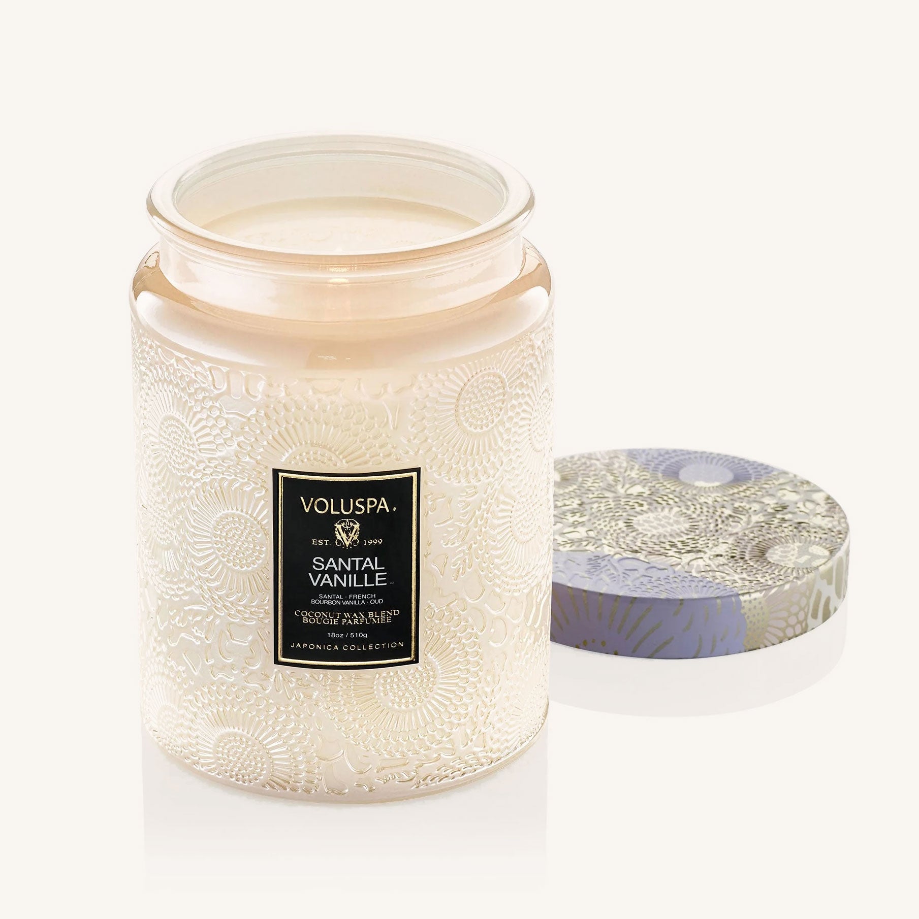 In front of a white background is a large light pink glass jar. The glass jar has a floral pattern all over it. In the middle is a black rectangular sticker with gold text at the top that reads ‘voluspa.’ Below is white text that reads ‘santal vanille.’ Leaning against the right side of the jar is a round tin lid. The lid has a light pink, silver and lavender flower pattern on it.