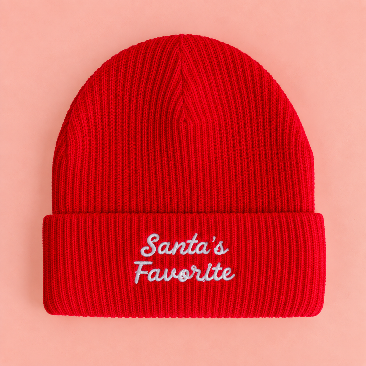 Product photo of a bright red knitted winter beanie, folded at the brim, with white cursive embroidery reading ‘Santa’s Favorite,’ centered against a pale pink backdrop