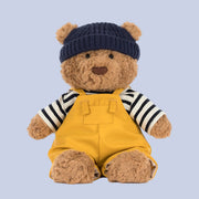Teddy bear wearing a navy hat, striped shirt, and yellow overalls on a white background